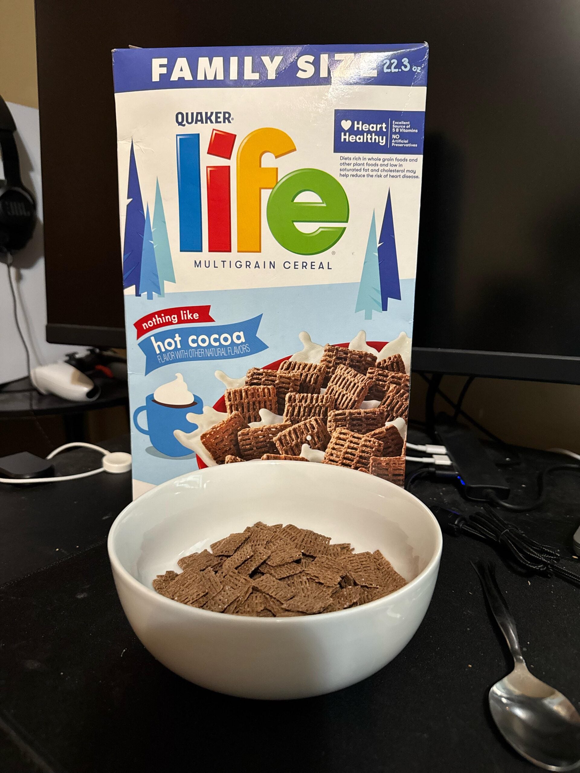 A box of Life Multigrain Cereal with a bowl of the chocolate cereal in front, sitting on a black desk near a spoon, headphones, and a computer monitor.