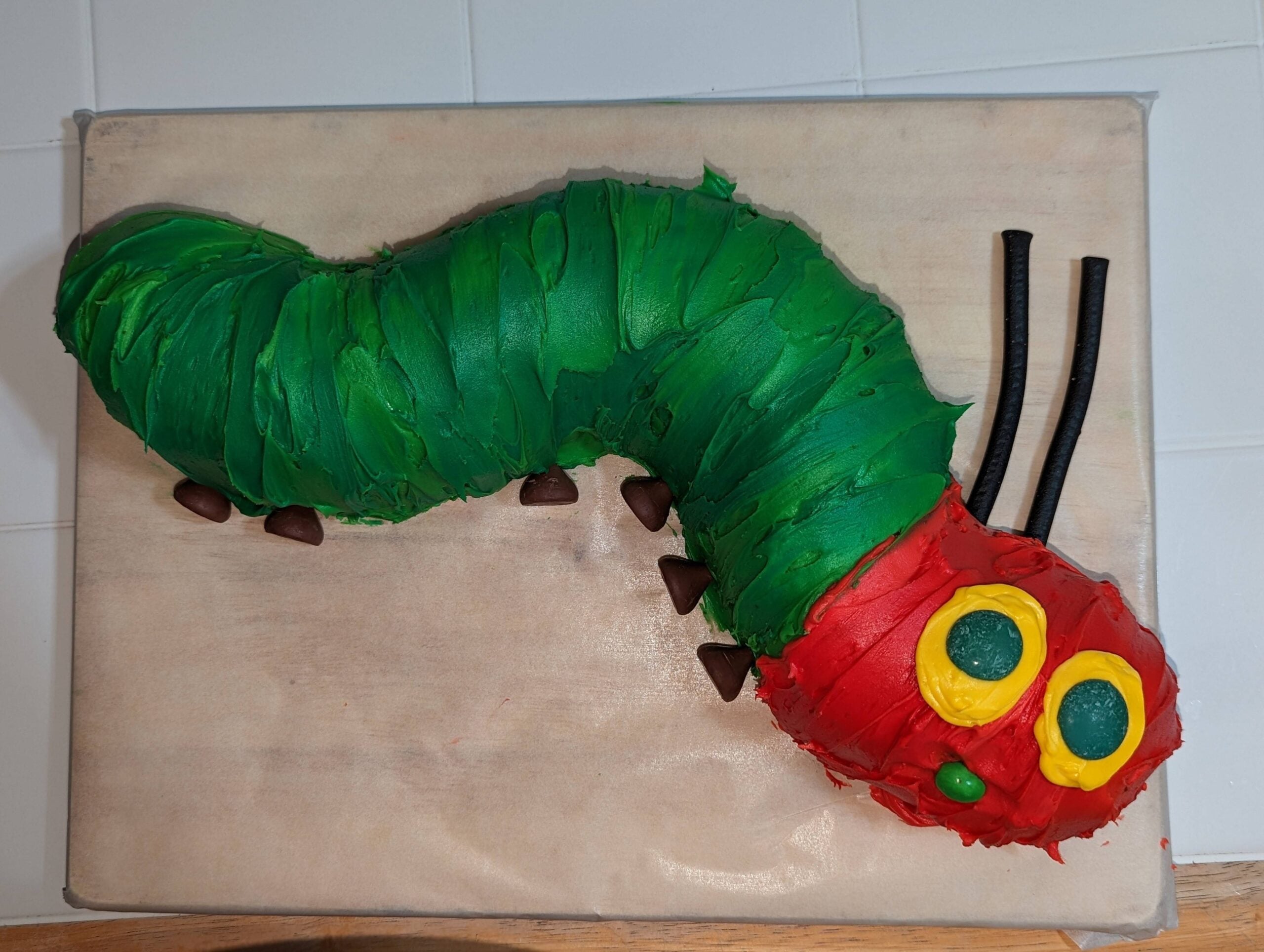 A cake shaped like a caterpillar, with green and red icing, yellow eyes, black licorice antennae, and chocolate feet, on a beige surface.
