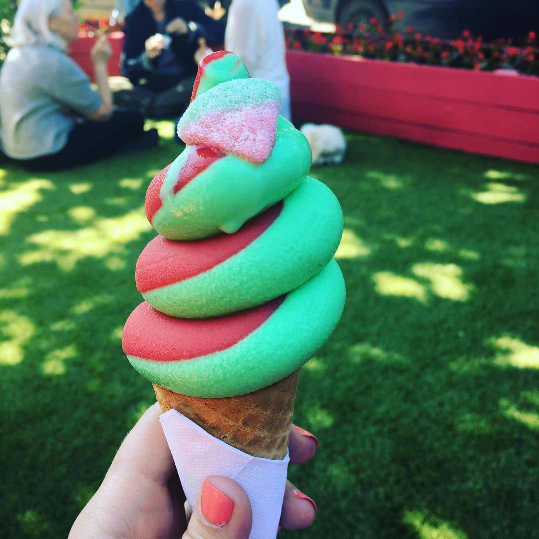 A hand holds a red and green swirled ice cream cone. In the background, people sit on grass in a sunny outdoor area with red planters.
