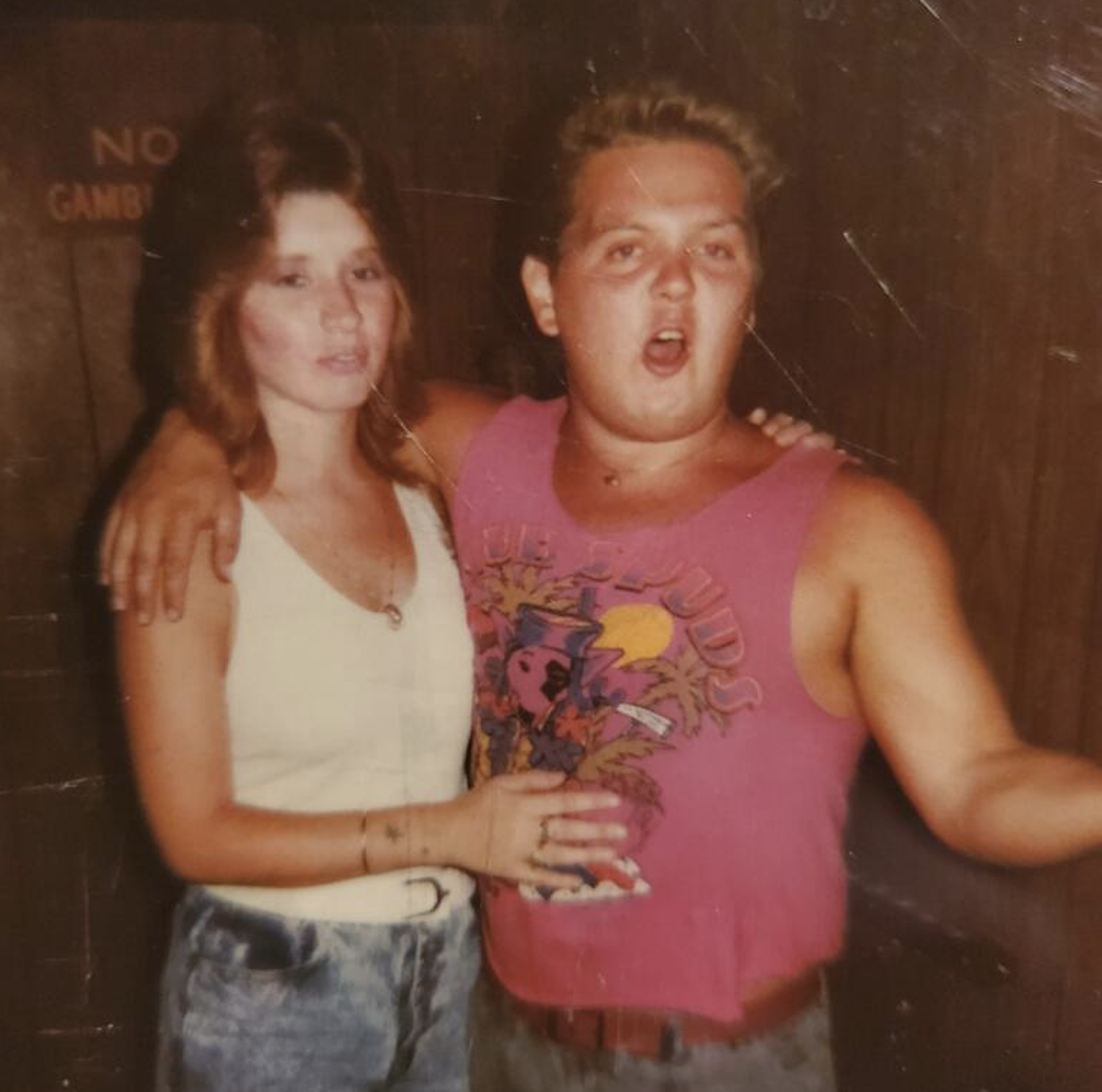 Two people pose together indoors. The woman wears a white tank top and jeans, while the man, in a bright pink graphic tank top, gestures with his mouth open. Both appear to be having fun.