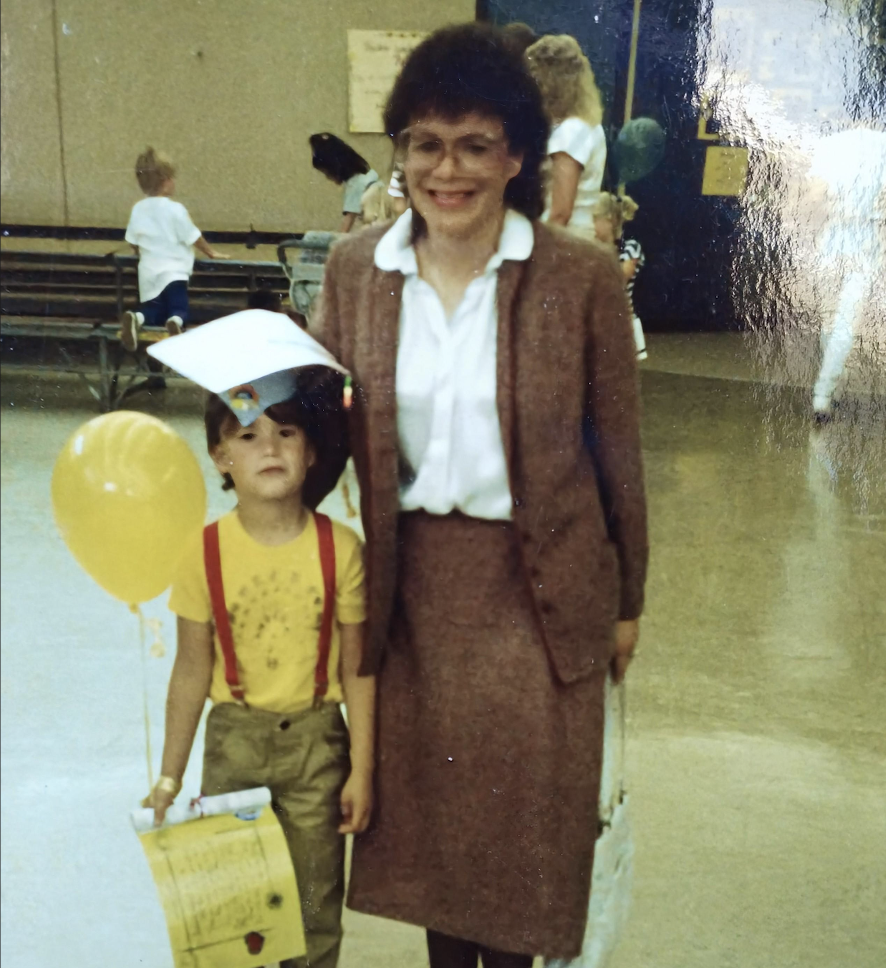A young child holding a balloon and certificate stands beside an adult woman in a suit, both smiling indoors during a school event.
