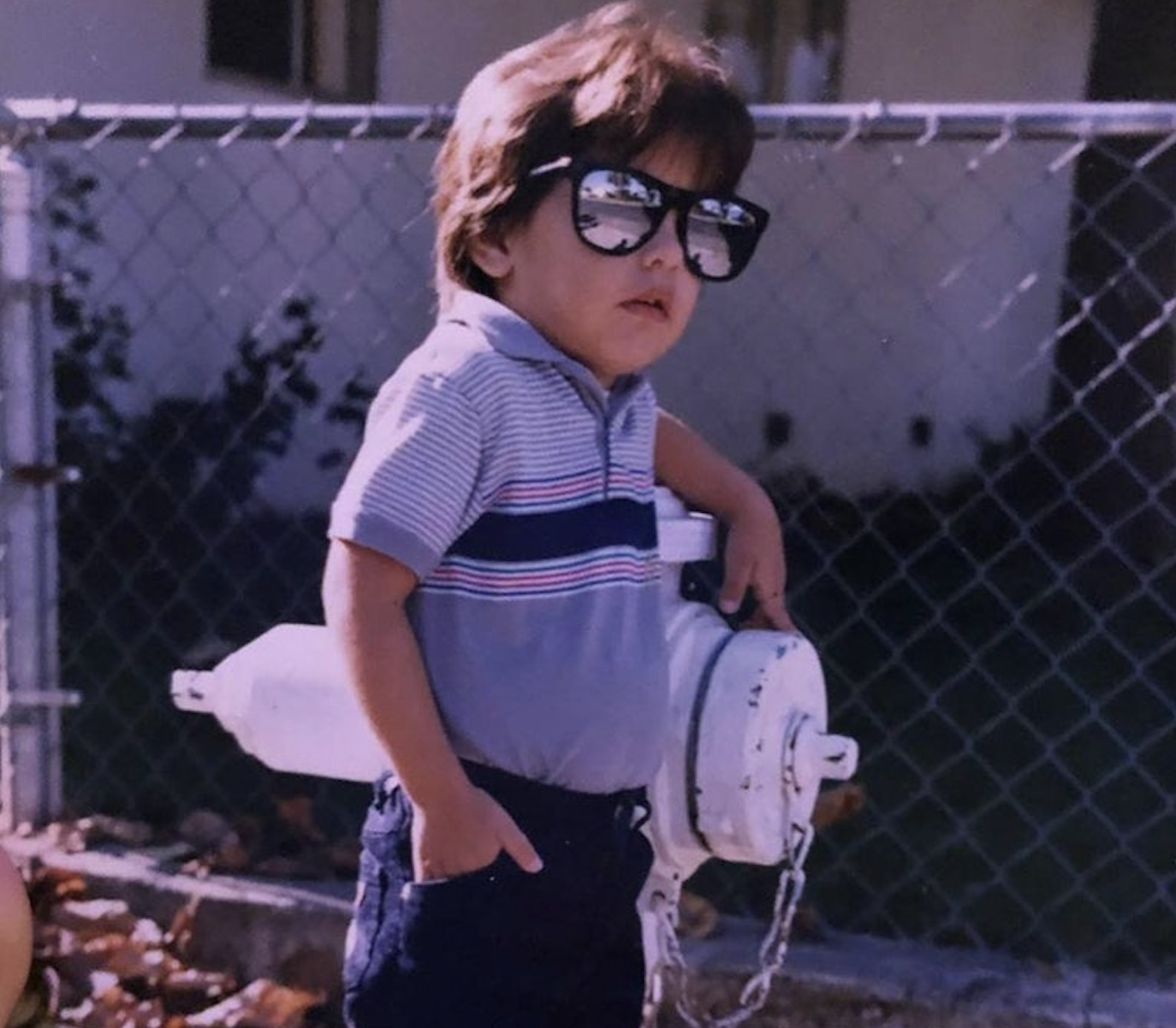 A young child in sunglasses stands outdoors by a fire hydrant, with one hand in their pocket, wearing a striped polo shirt and dark pants. There is a chain-link fence and building in the background.