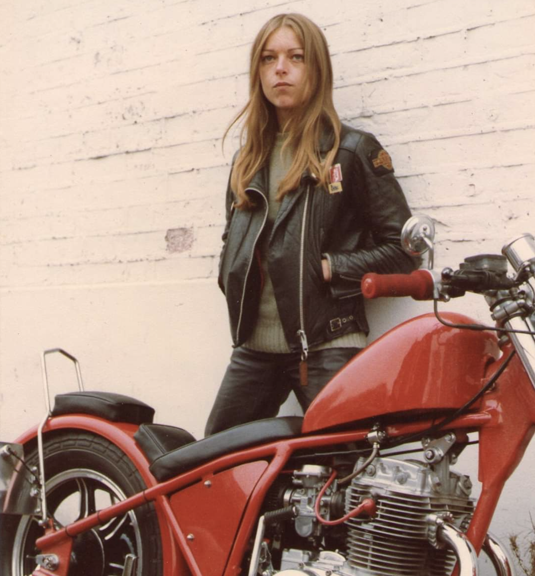 A woman with long hair in a leather jacket stands against a white brick wall next to a red motorcycle, looking at the camera with a serious expression.