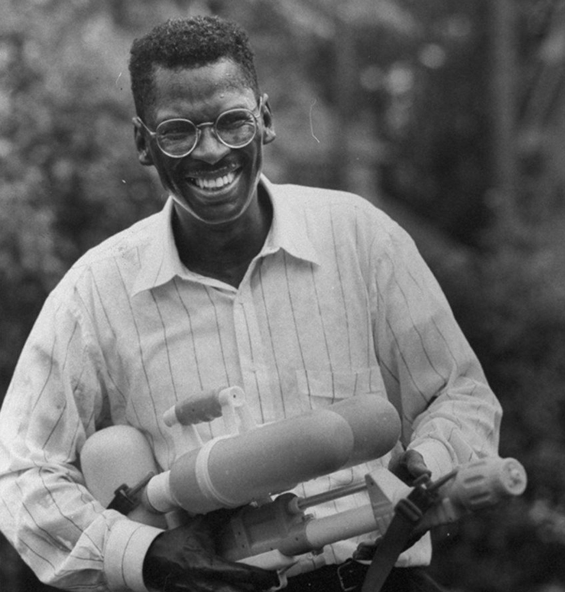 A smiling man wearing glasses and a striped shirt holds a large toy water gun outdoors.