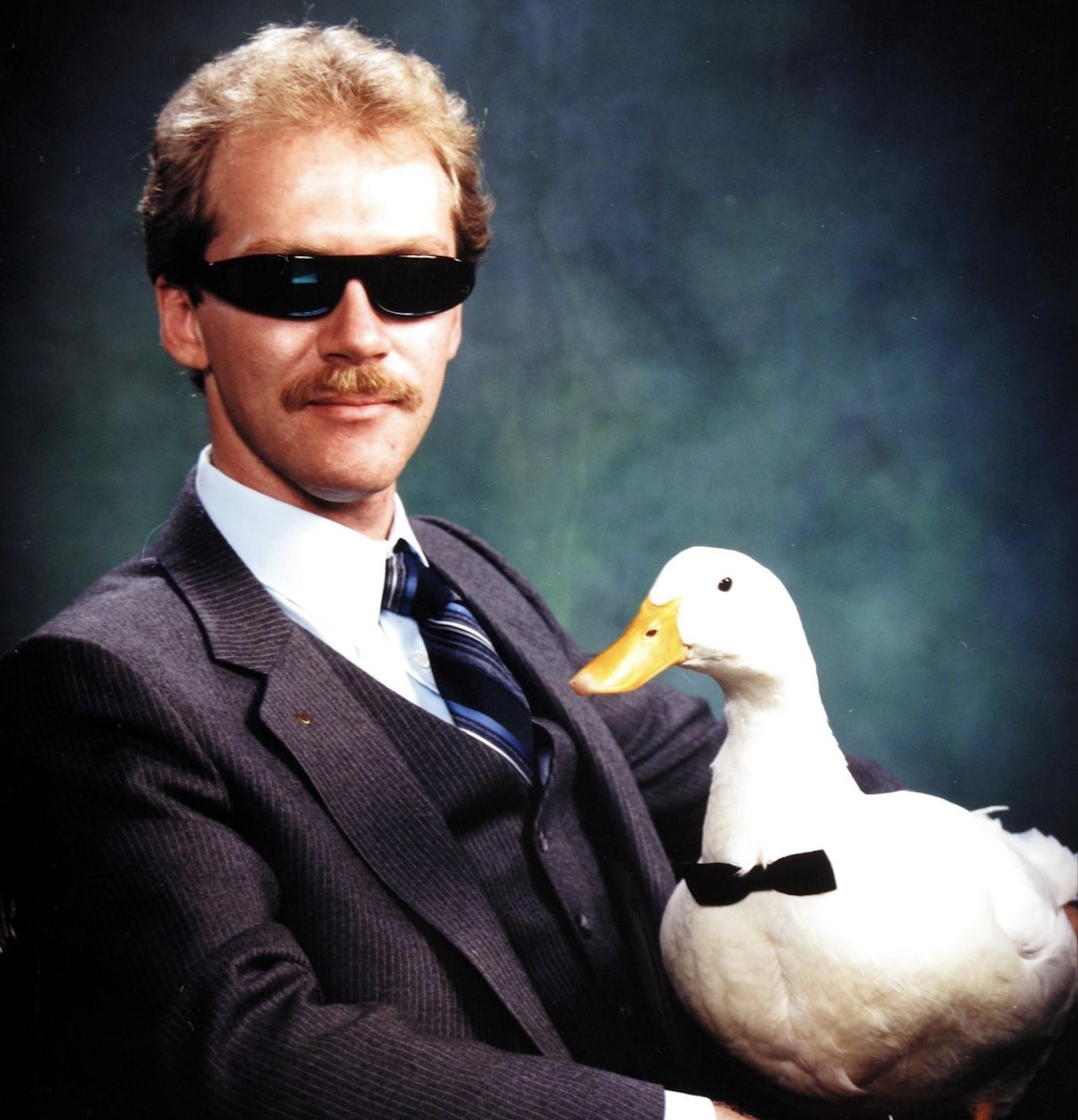 A man in a suit and sunglasses poses with a white duck wearing a black bowtie against a dark, gradient studio backdrop.