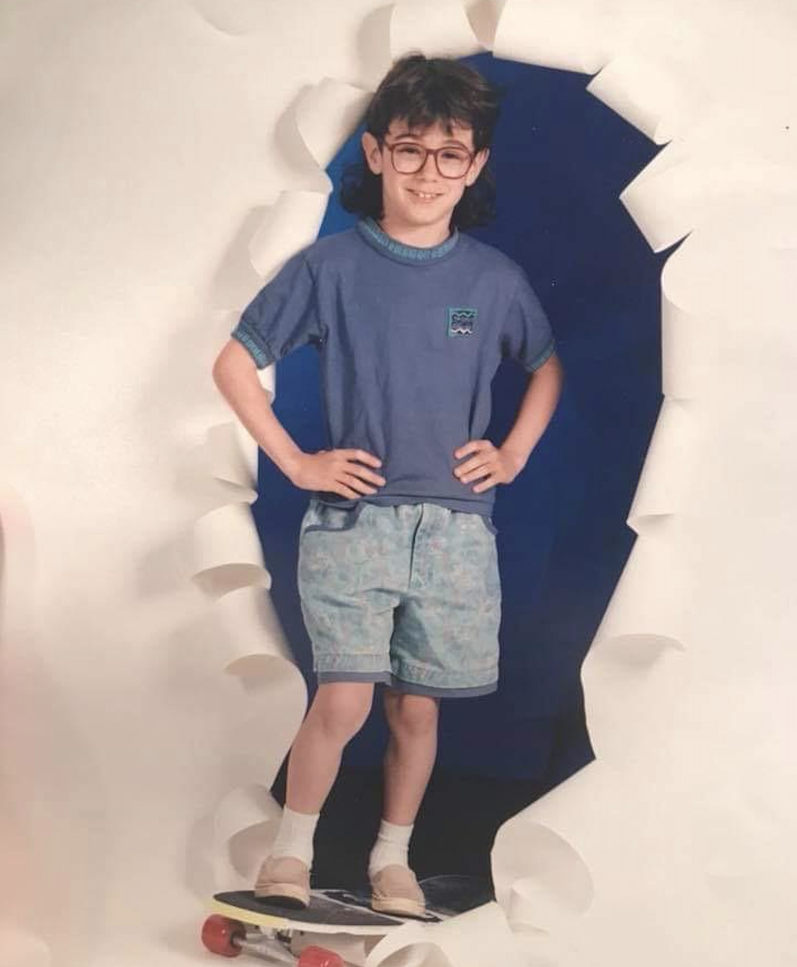 A child with glasses stands on a skateboard, posing confidently in front of a backdrop designed to look like torn paper. The child wears a blue shirt, patterned shorts, and white socks with shoes.