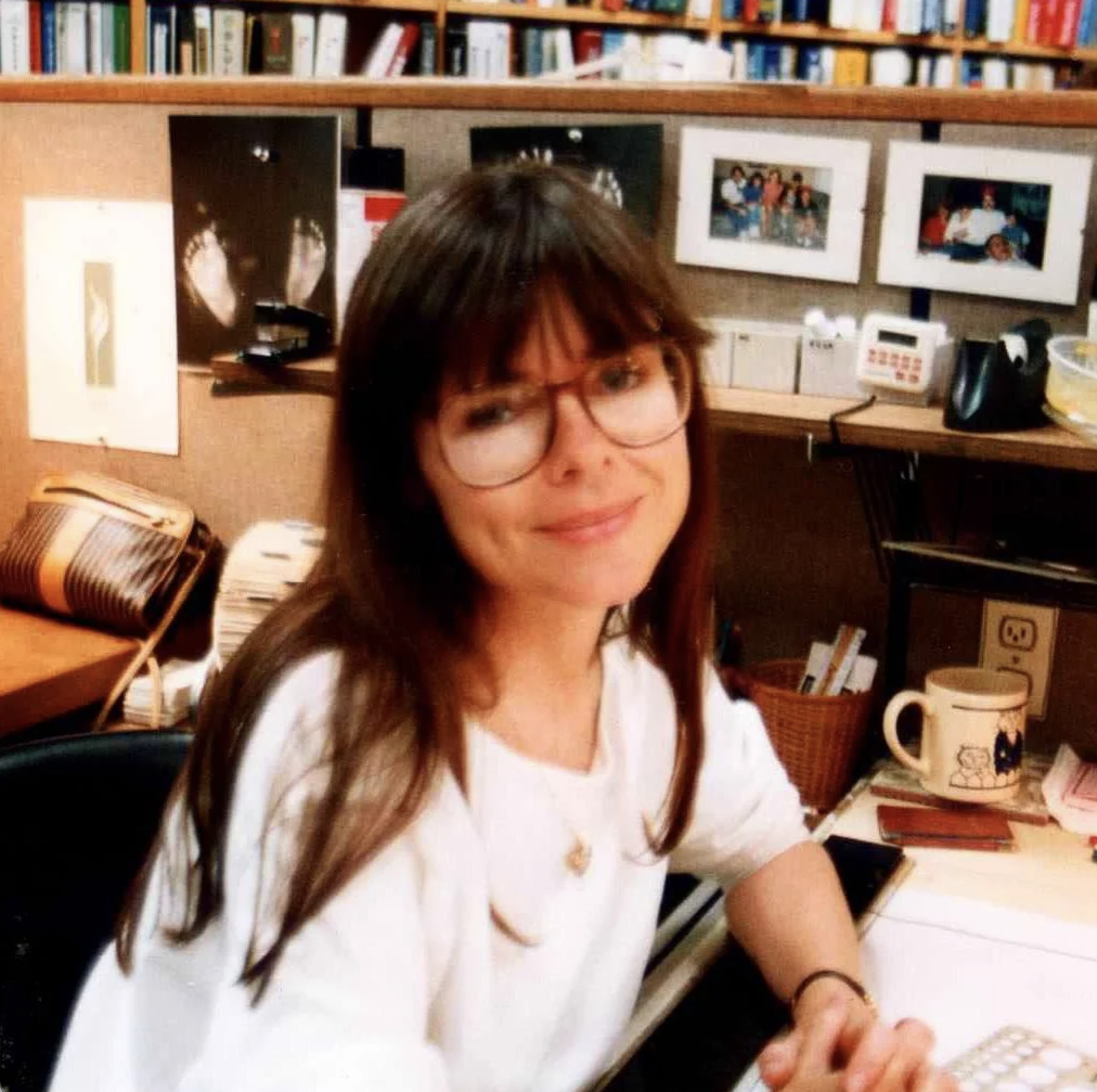 A woman with long brown hair and large glasses smiles at her desk in an office, surrounded by photos, books, a phone, and a mug.