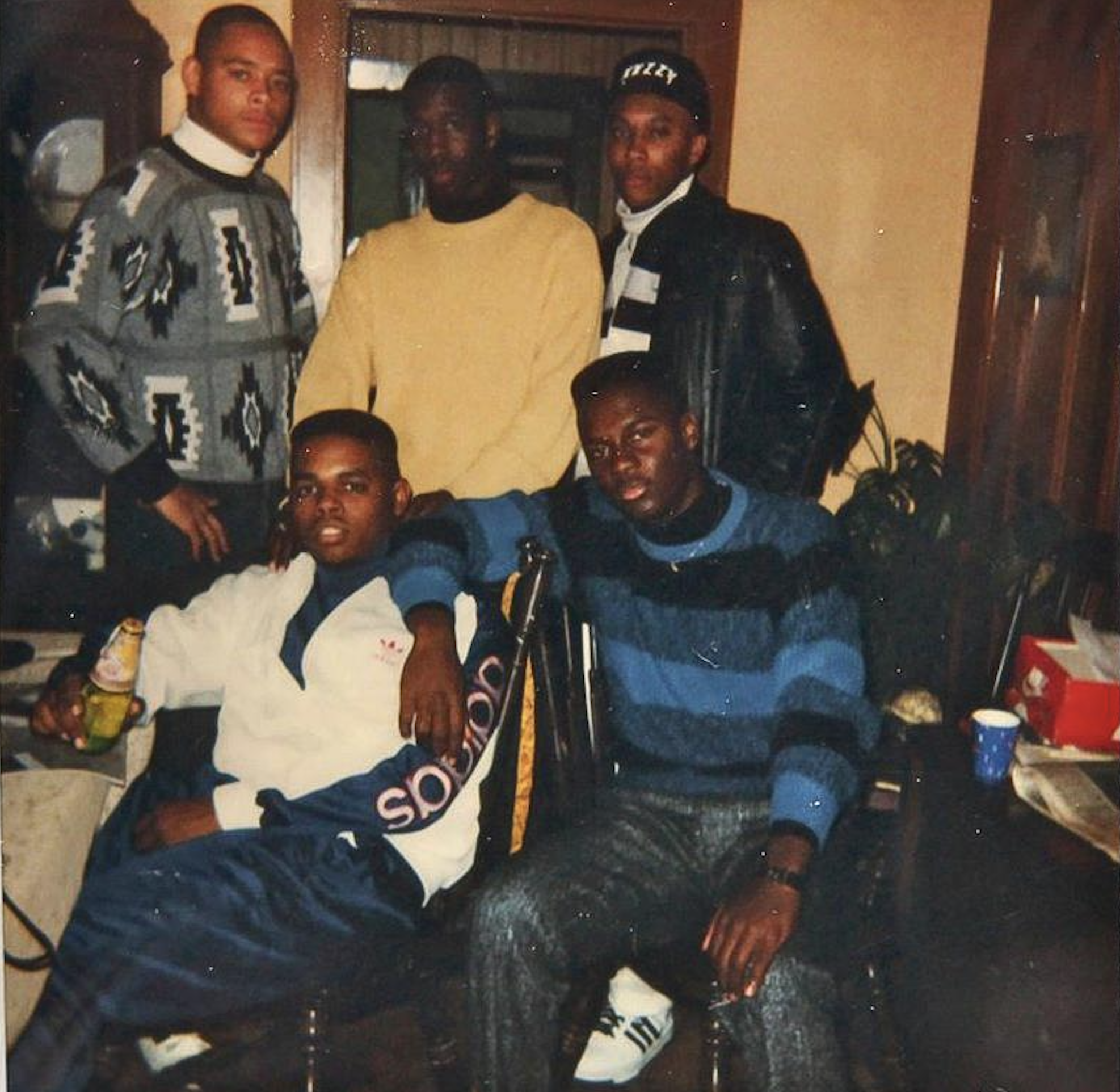 Five young men indoors pose for a photo. Three stand in the back, and two sit in front. They wear sweaters, jackets, and tracksuits. The room has wood trim, plants, and a cluttered table with drinks and paper items.