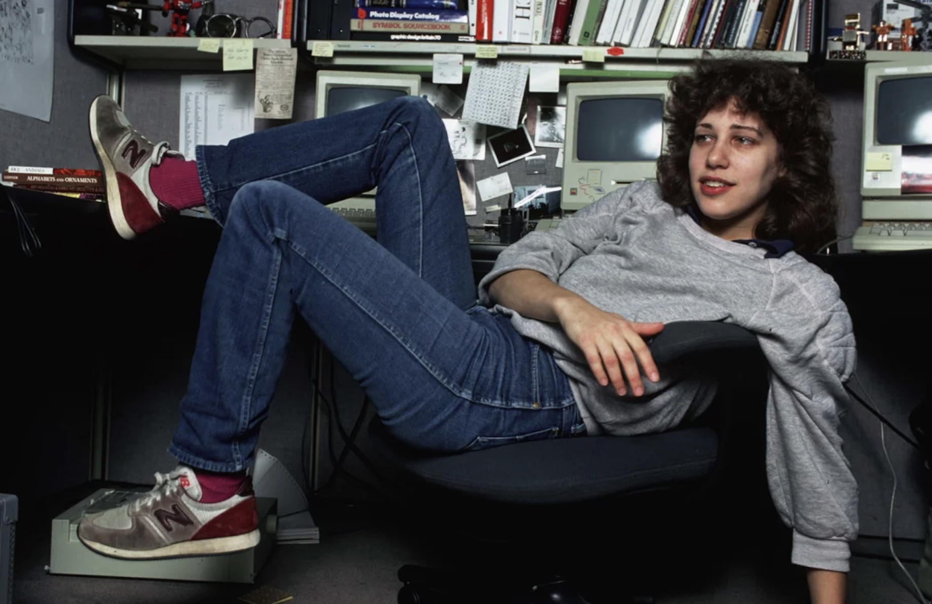 A person in jeans and sneakers lounges on an office chair with their legs propped on a desk, surrounded by vintage computers, books, and office supplies.