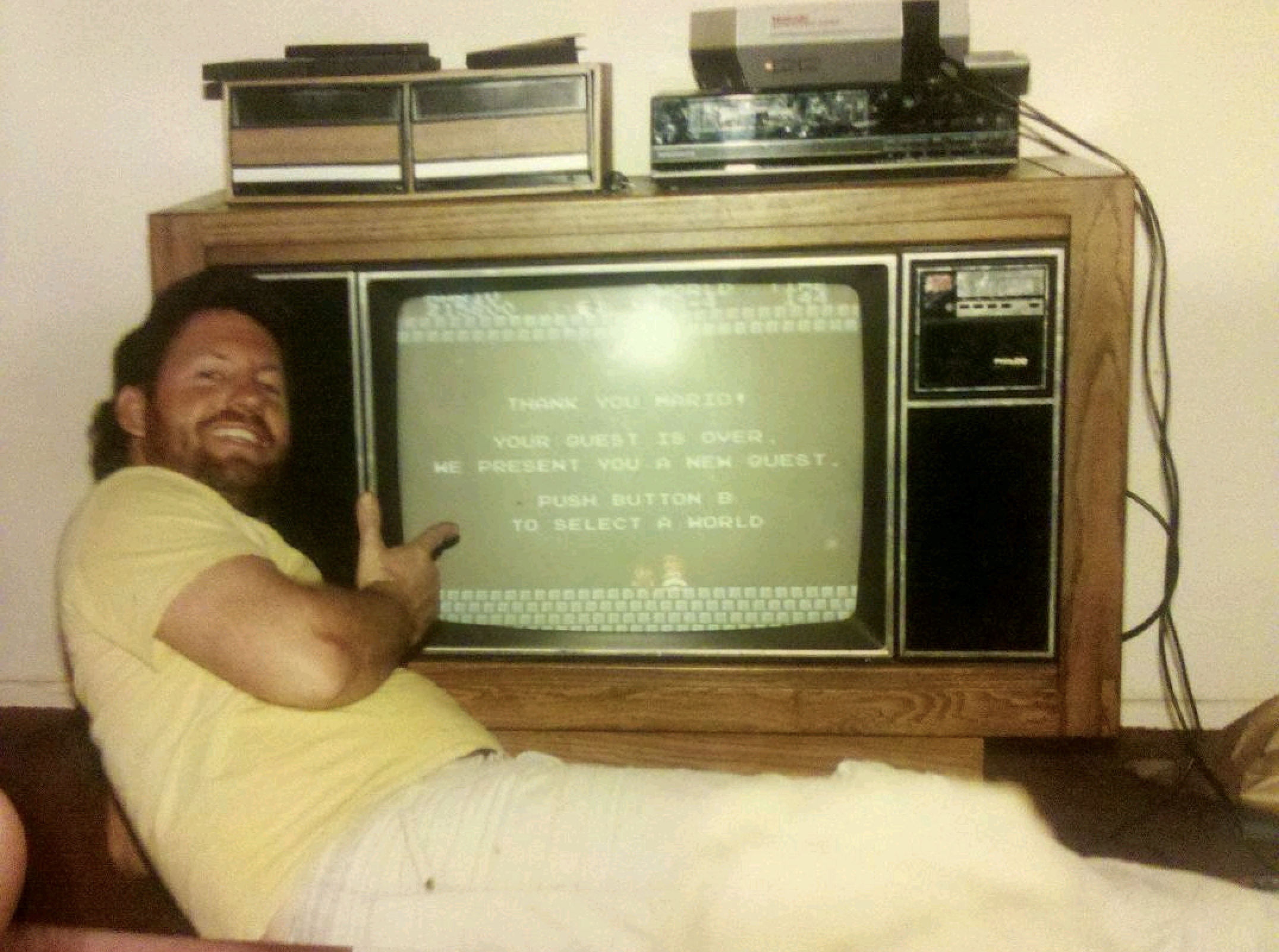 A man in a yellow shirt smiles and points at an old TV displaying a congratulatory message from a video game. A vintage game console and VCR sit on the wooden TV stand.