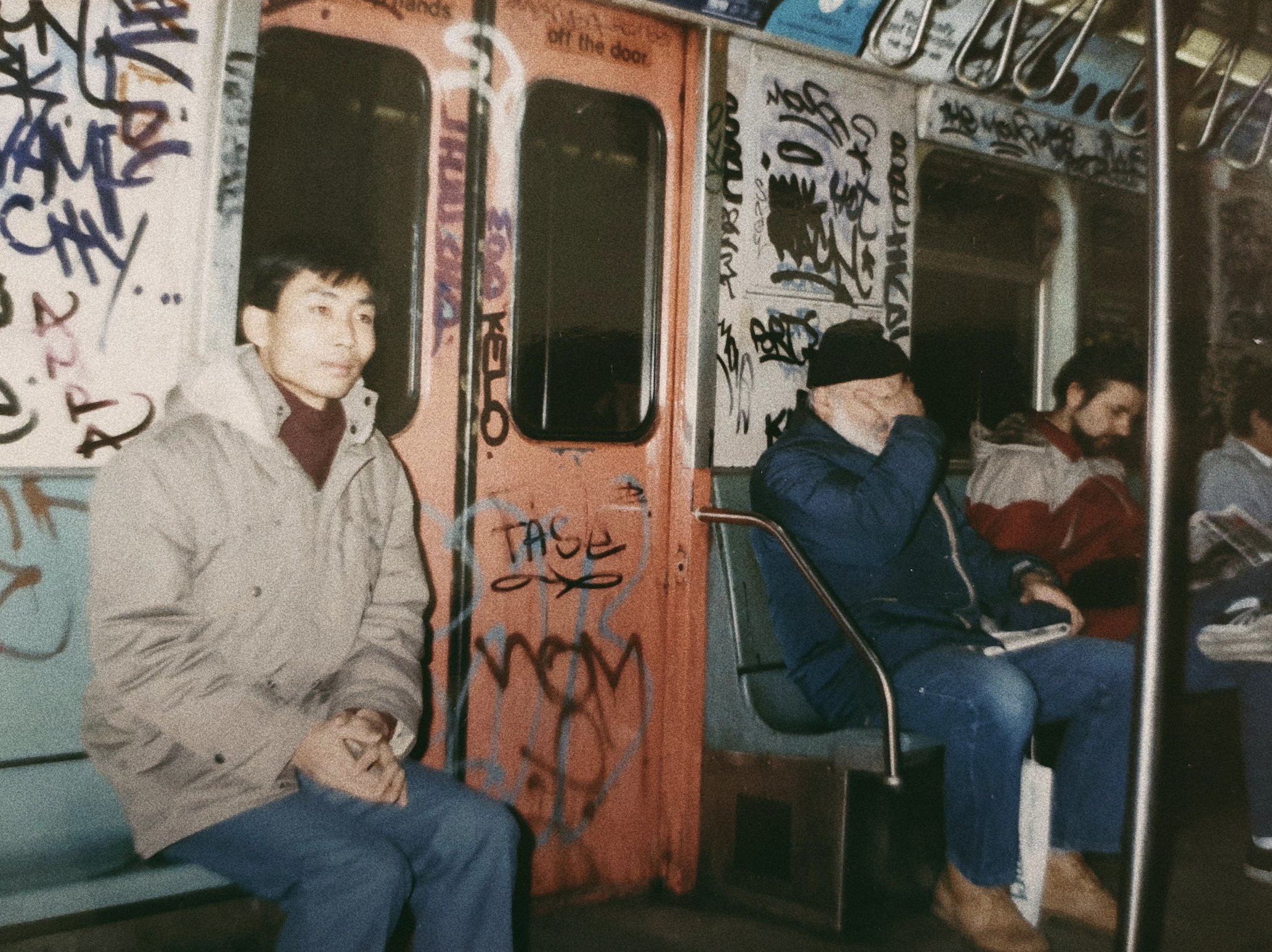 Three men sit on a graffiti-covered subway train; one faces forward, another covers his face, and the third reads. The train is old with worn seats and tag-covered doors and walls.