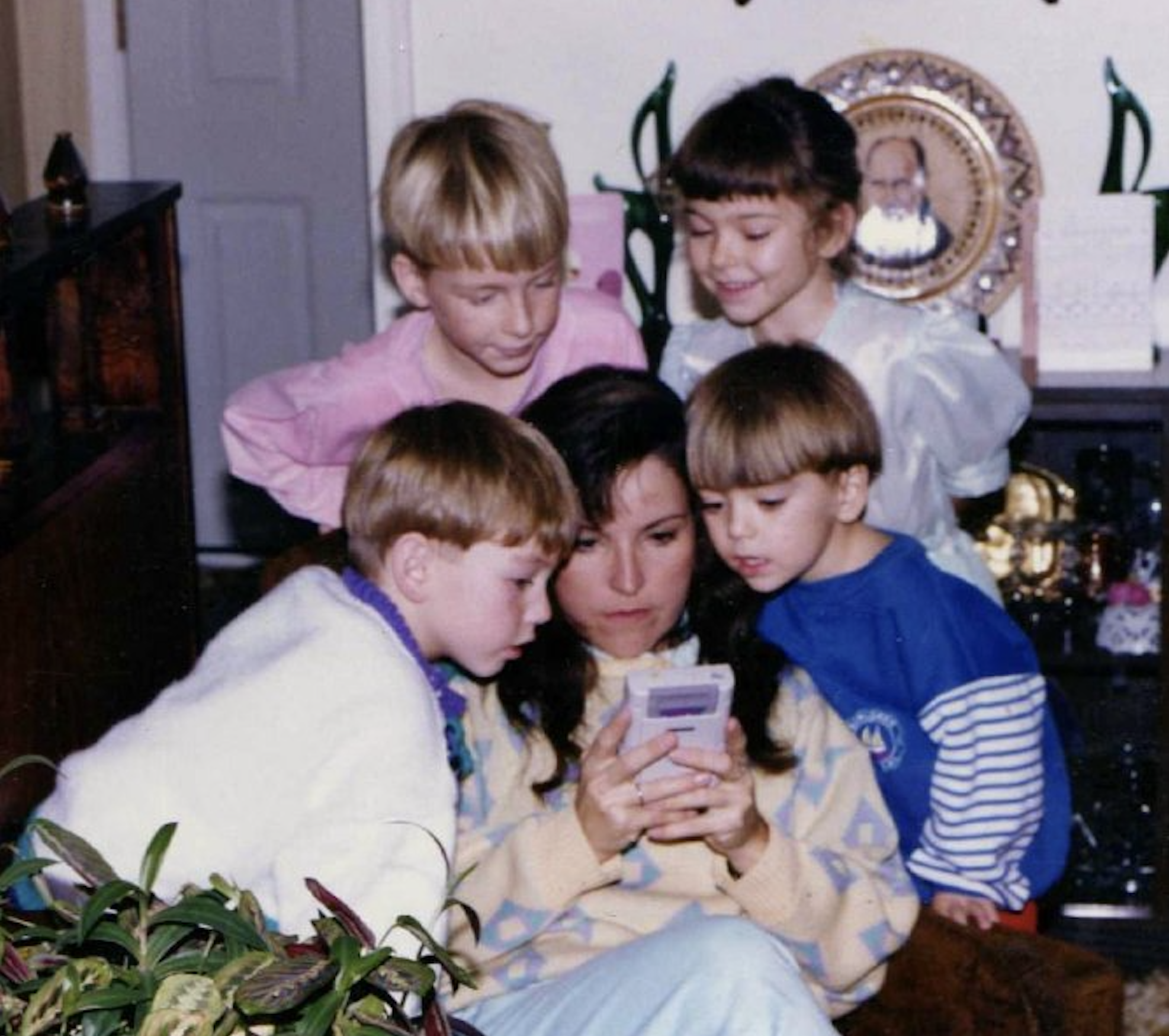 A woman sits on a chair playing a handheld game console, surrounded by four young children who eagerly watch her play. There’s a plant in the foreground and framed photos in the background.
