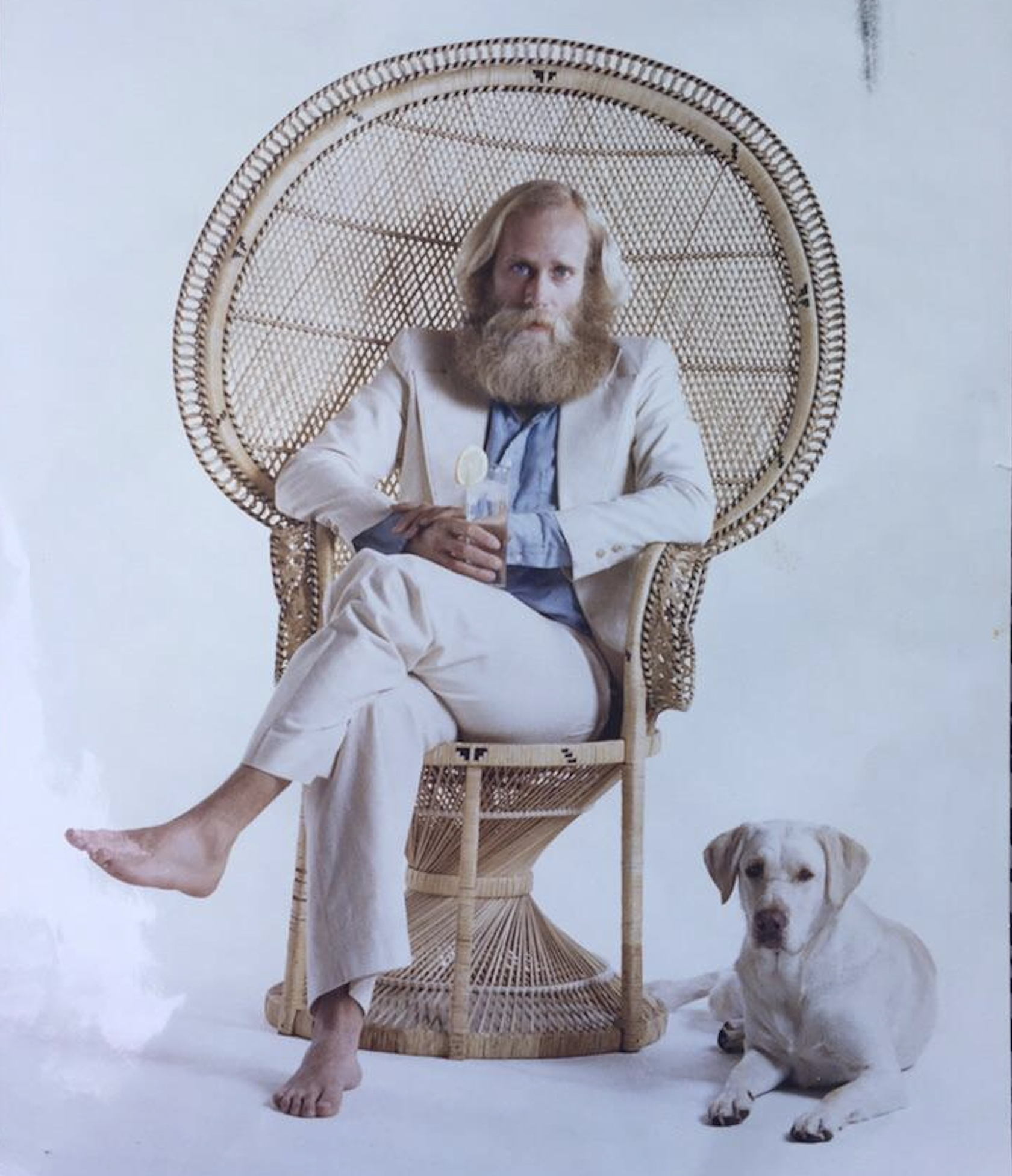 A barefoot man with a long blond beard sits in a peacock chair holding a drink, dressed in a light suit. A Labrador dog lies on the floor by his feet. The background is plain and white.