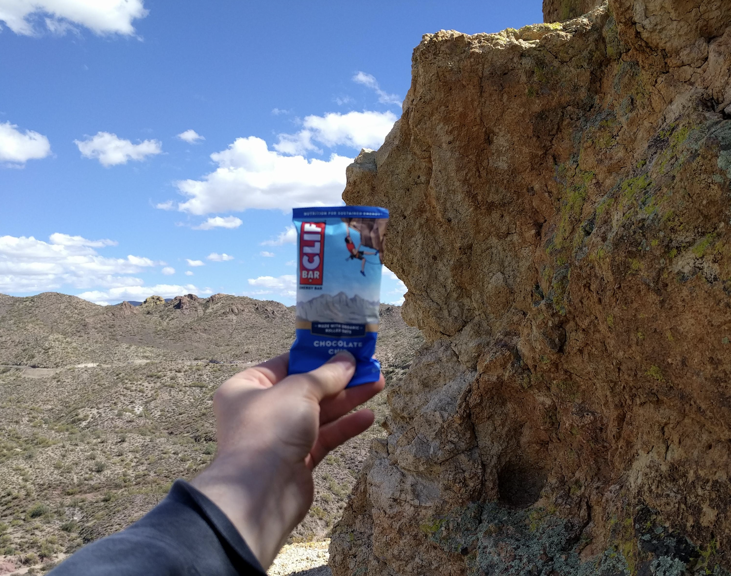 A hand holds a blue-wrapped Clif Bar in front of a rocky cliff, with a mountainous desert landscape and partly cloudy sky in the background.