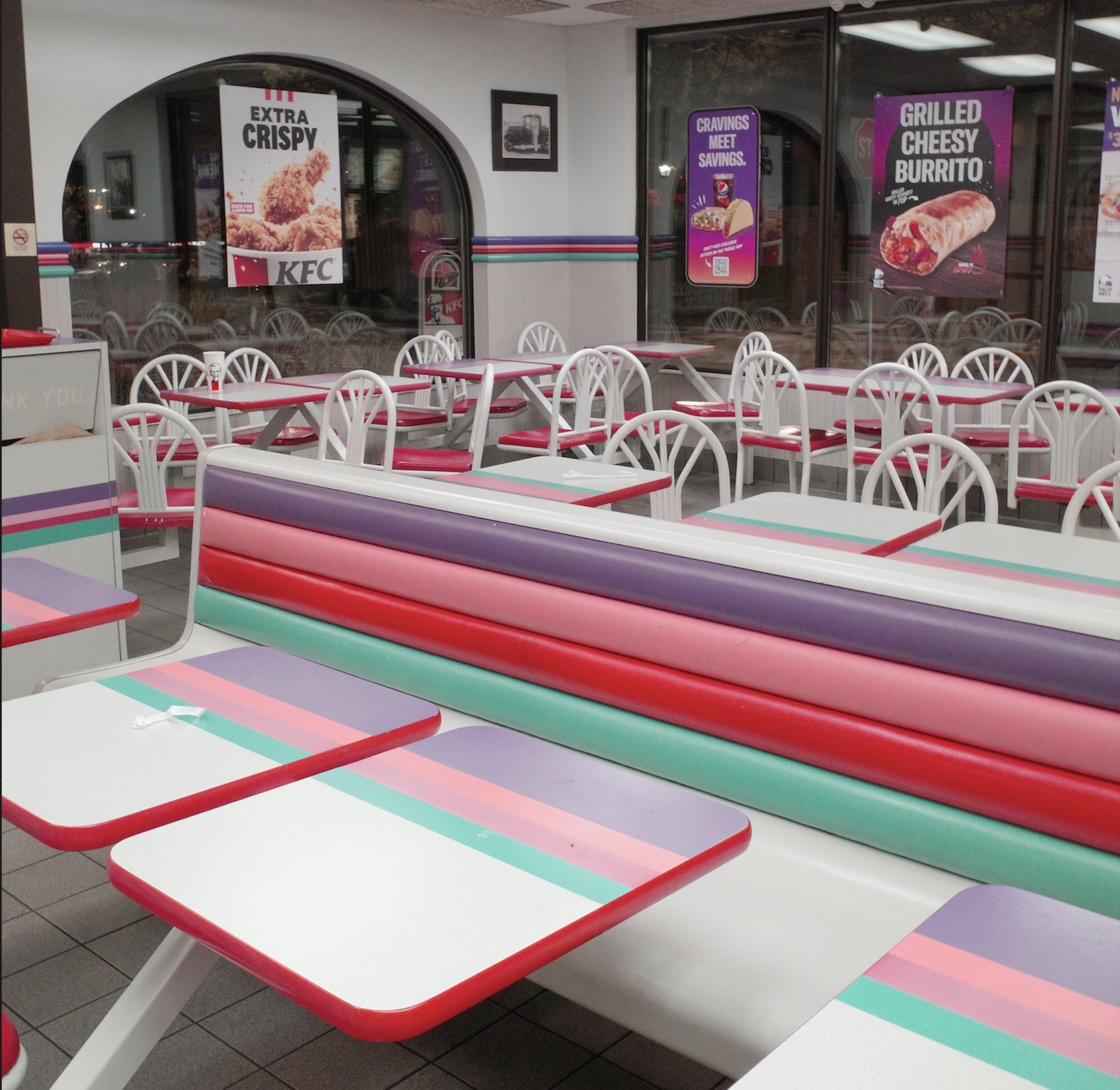 Empty fast-food restaurant interior with colorful striped benches and white chairs. Brightly lit space, three large windows display posters advertising KFC, Cinnabon Delights, and Grilled Cheesy Burrito.