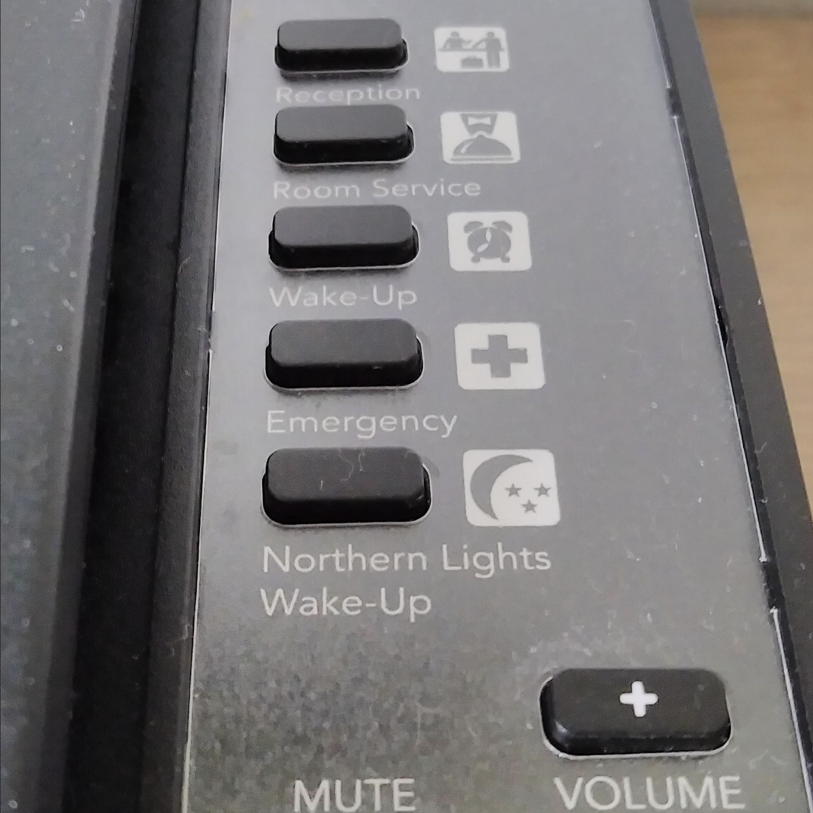 Close-up of black buttons on a device labeled: Reception, Room Service, Wake-Up, Emergency, Northern Lights Wake-Up; icons appear next to each label. MUTE and VOLUME buttons are partially visible below.