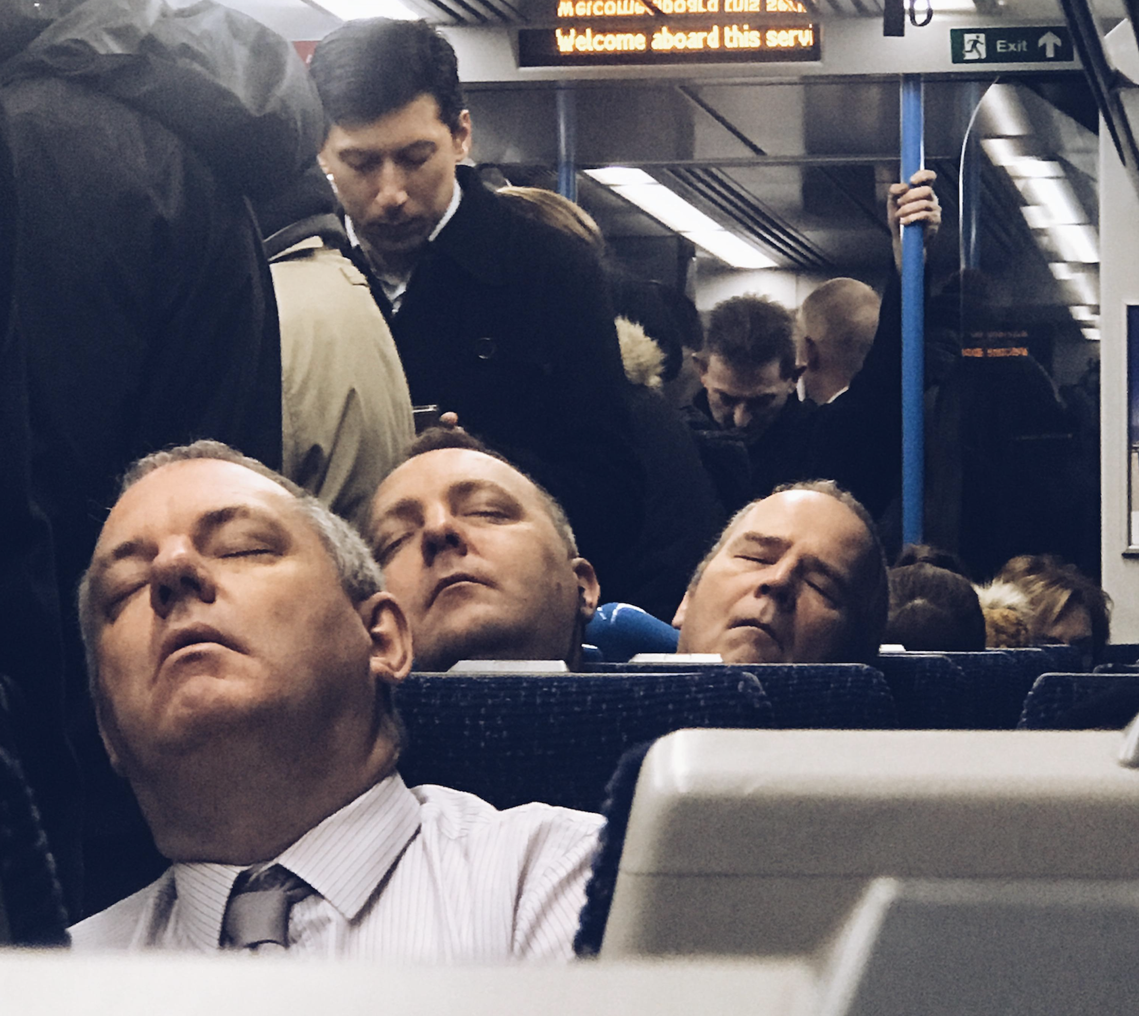 Three men in suits sit in a row on a crowded train, all sleeping with heads tilted back. Other passengers stand and look at phones. The scene is dimly lit and the train appears full.