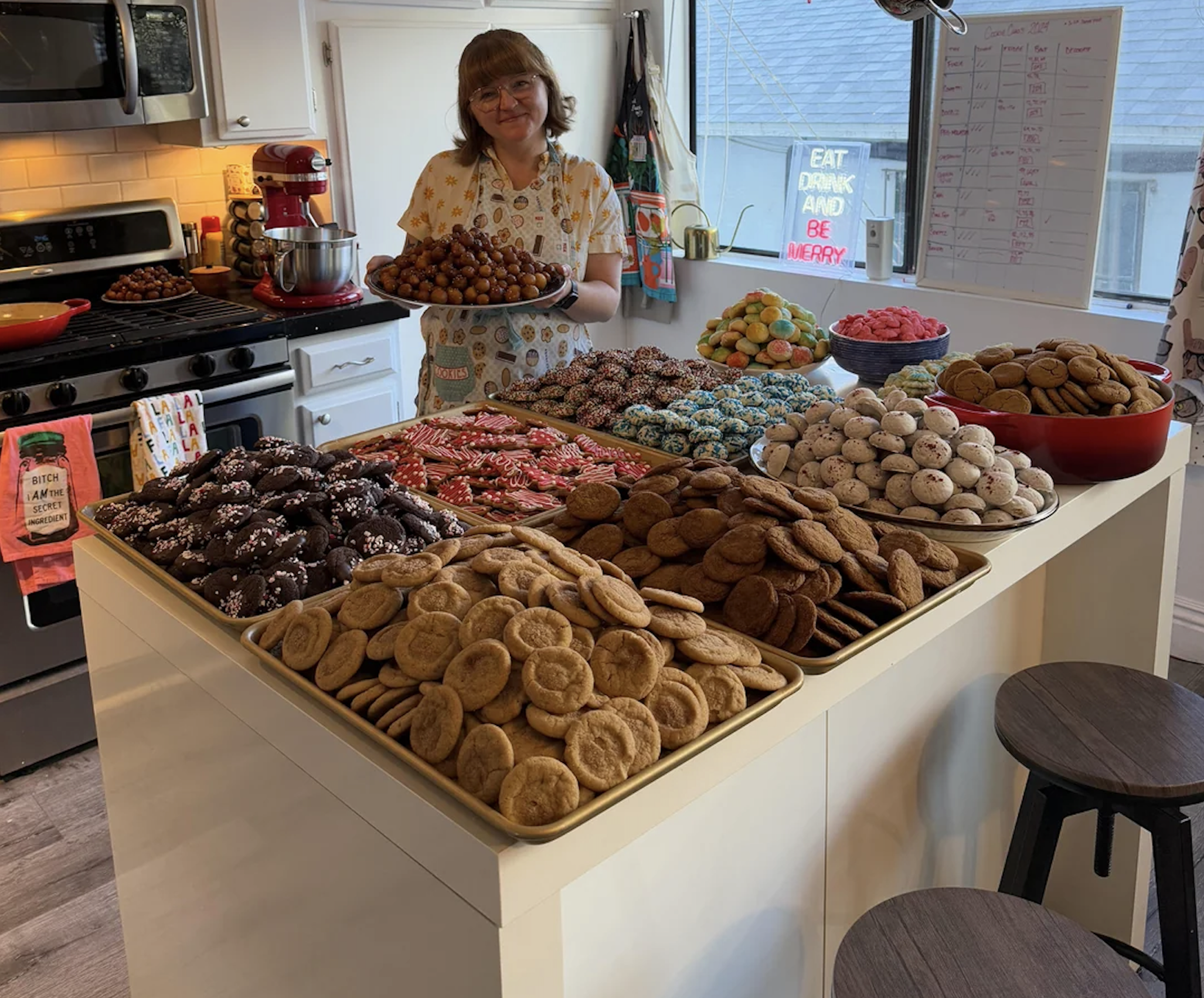 A person stands in a kitchen holding a plate of treats, with a large table covered in assorted cookies and sweets. The kitchen is bright, and a colorful sign on the window reads, “EAT DRINK AND BE MERRY.”