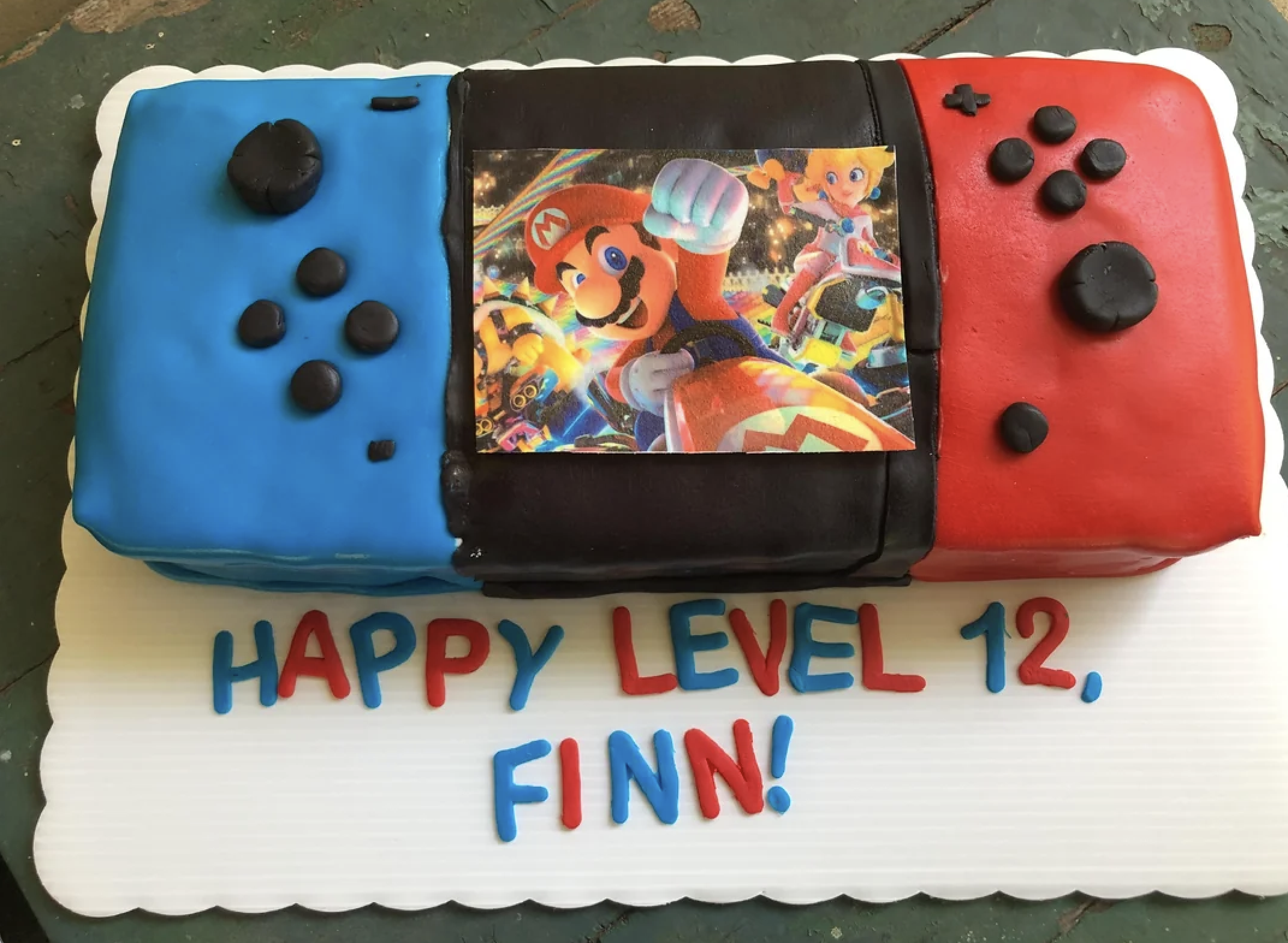 Rectangular cake designed like a Nintendo Switch with blue and red controllers, featuring an edible Mario image in the center. Text on the board reads: “HAPPY LEVEL 12, FINN!” in colorful letters.