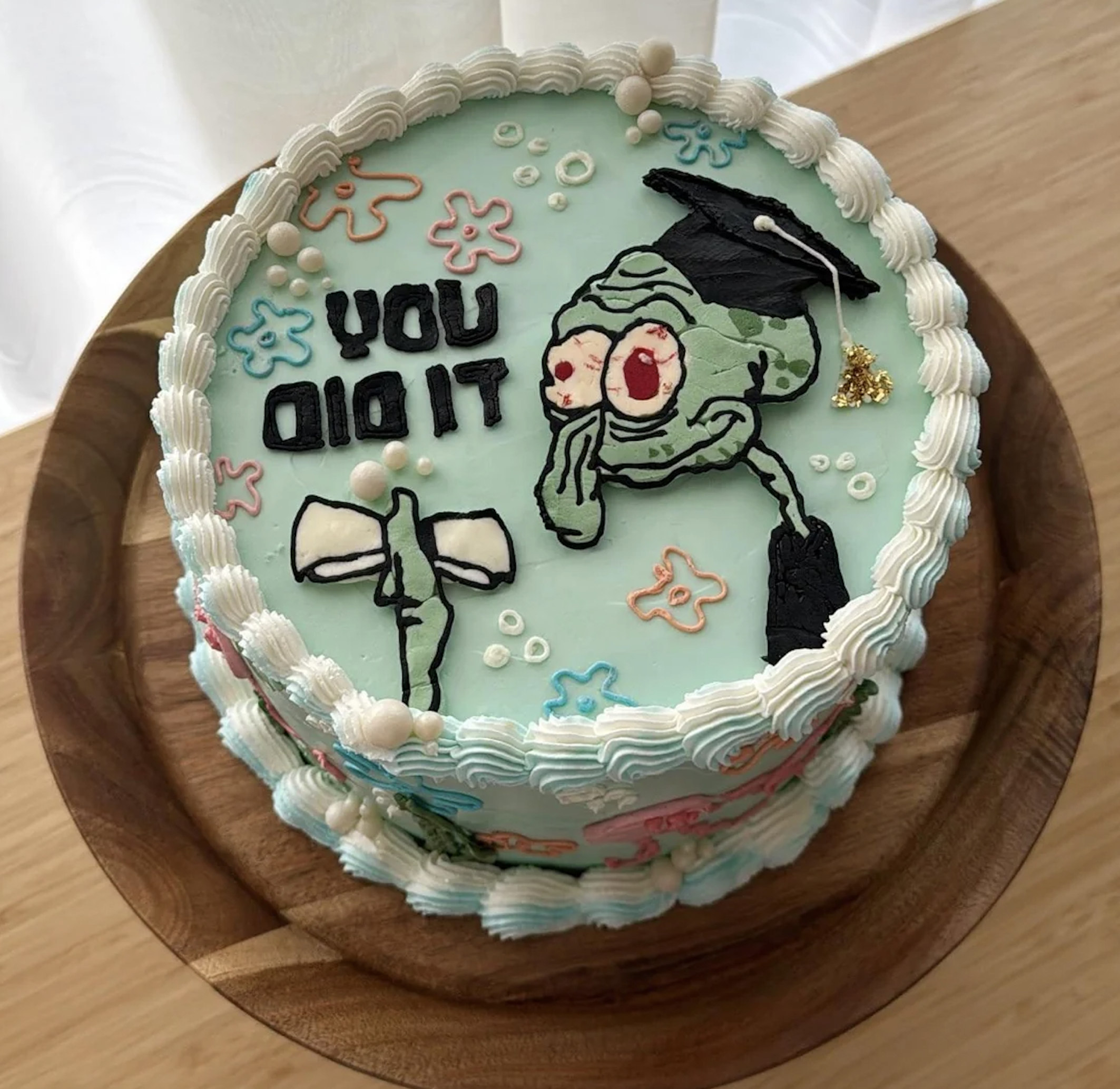 A round cake with Squidward in a graduation cap, holding a diploma. Text reads “YOU DID IT.” The cake has blue icing, scalloped edges, and white pearl decorations.