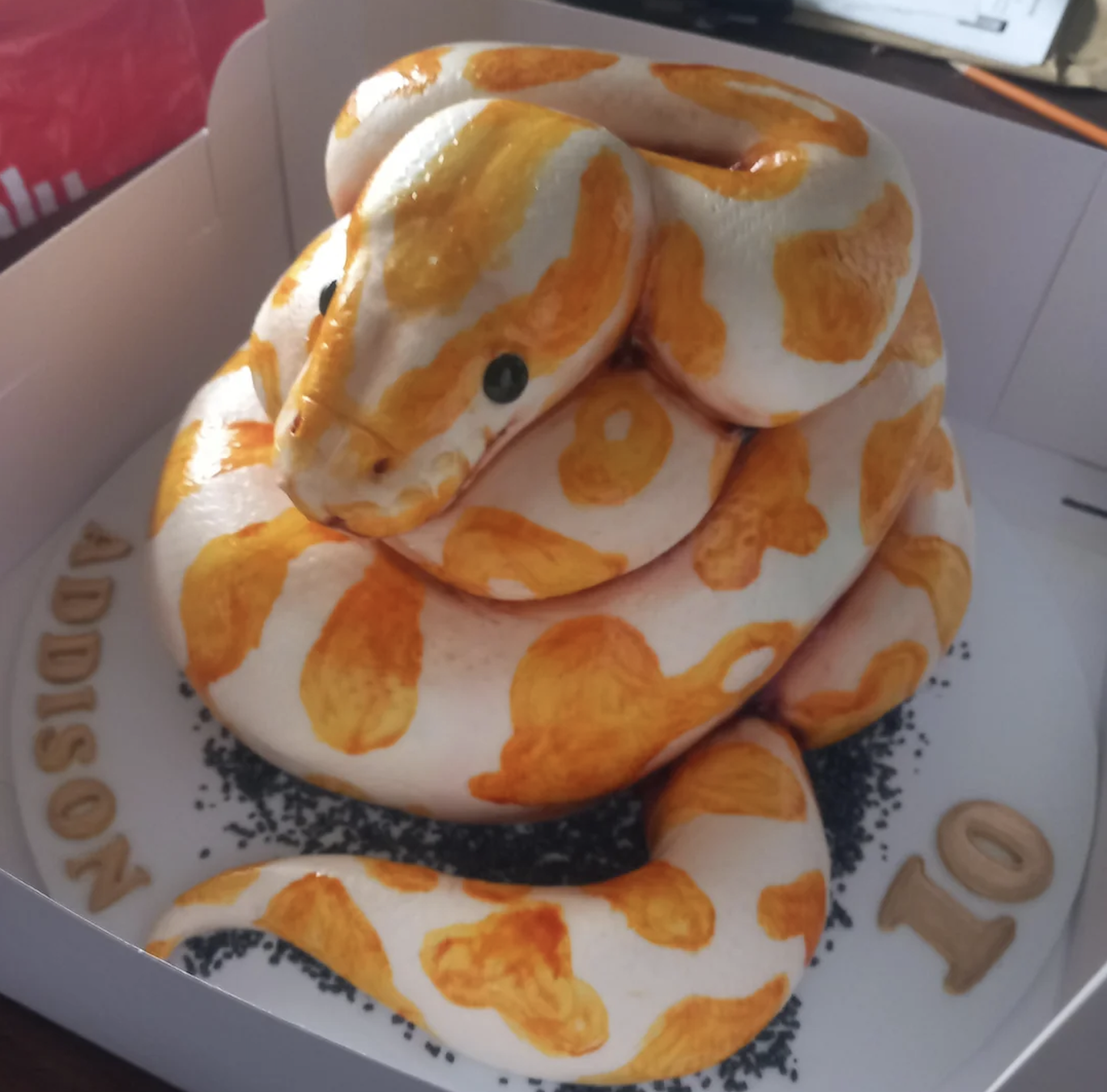 A realistic cake shaped like an orange and white snake sits in a box. The base says “ADDISON 10” with sprinkles around the cake.