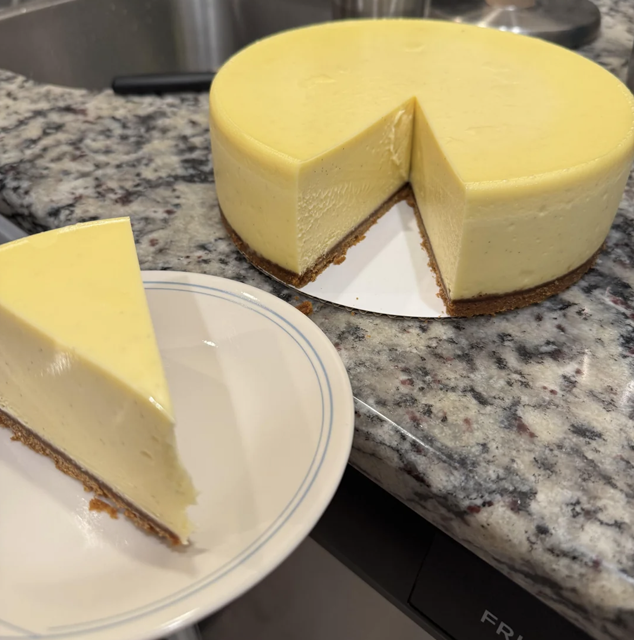A whole cheesecake with a slice removed sits on a countertop. The removed slice is placed on a white plate with a blue rim, showing a graham cracker crust and creamy filling.