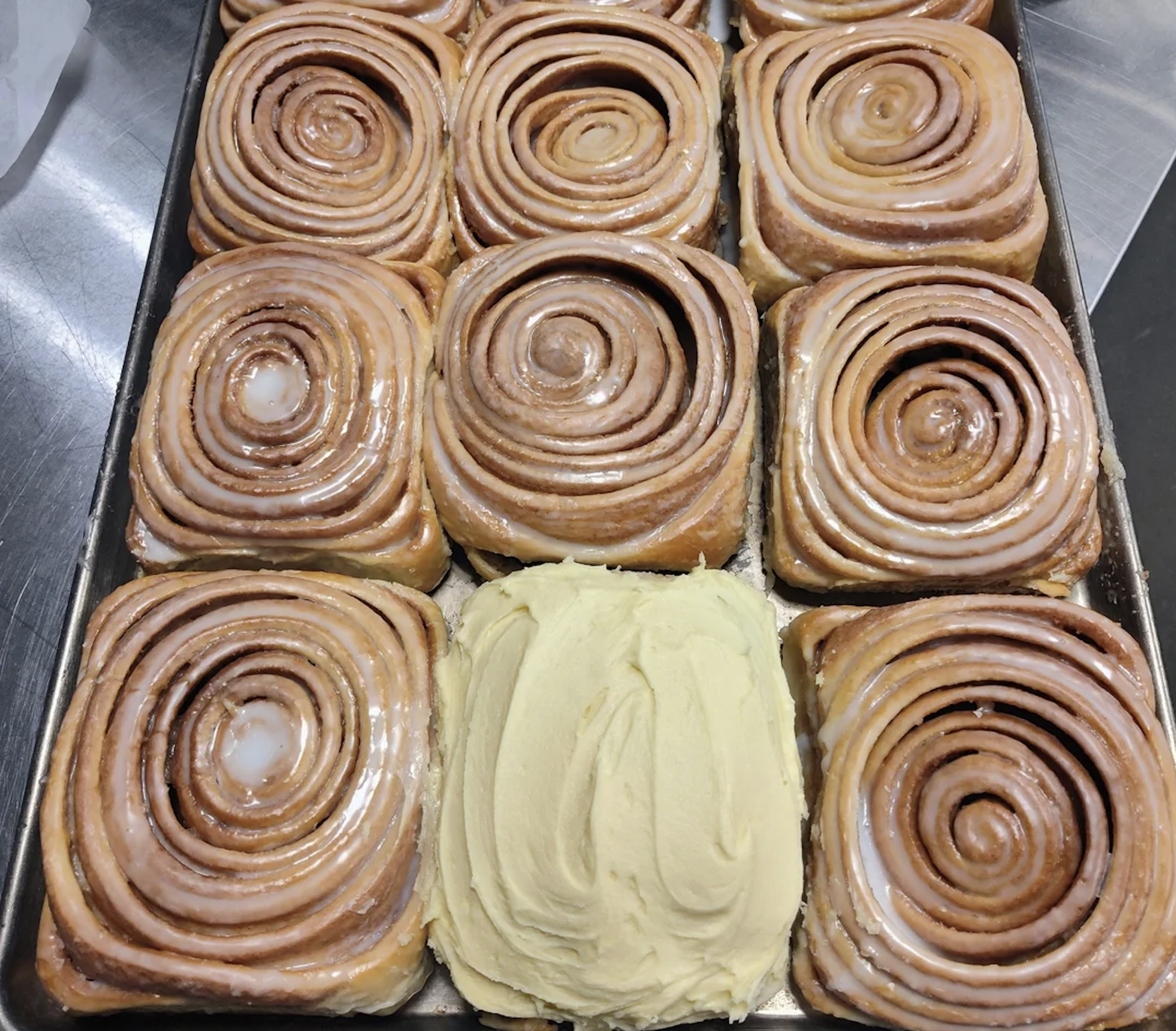 A tray of square cinnamon rolls with icing, except for one roll in the center bottom row, which is unfrosted and topped with swirled cream-colored frosting.