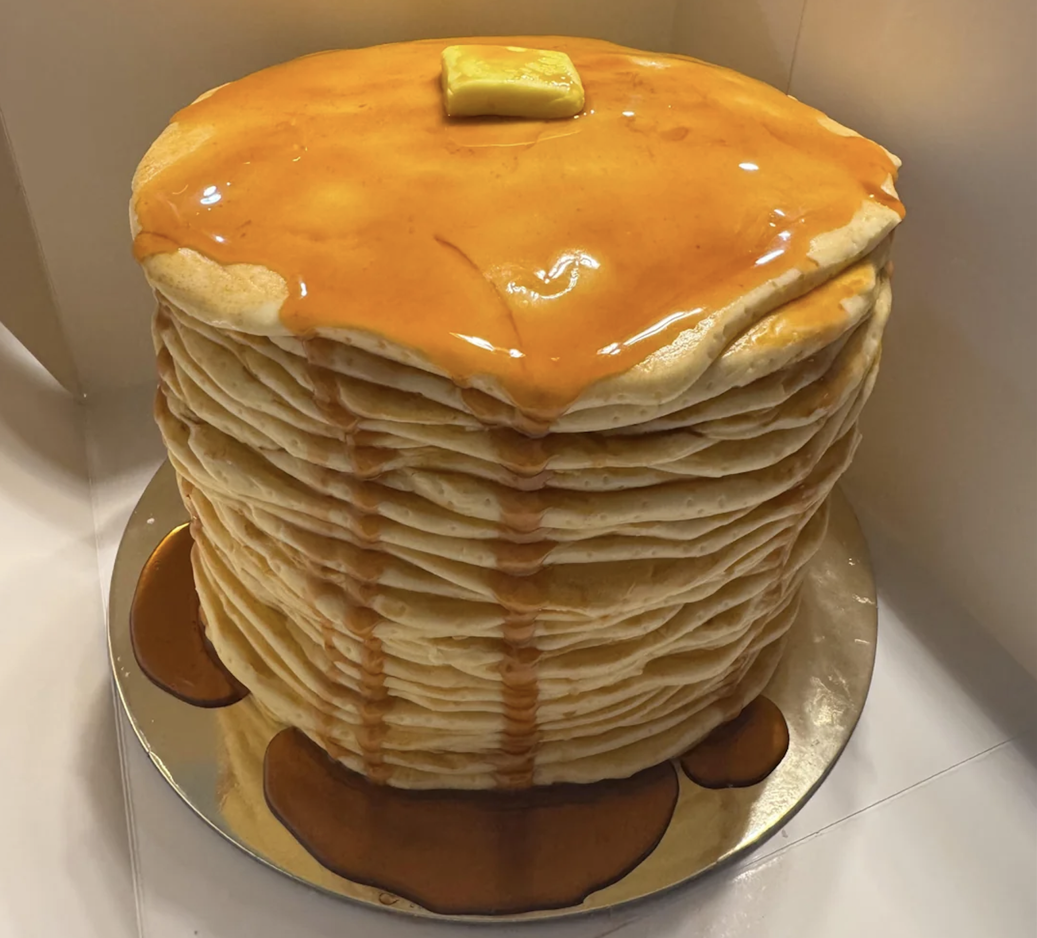 A tall, realistic cake decorated to look like a stacked tower of pancakes with syrup and a pat of butter on top, sitting on a reflective board. Syrup drips down the “pancakes” and pools at the base.