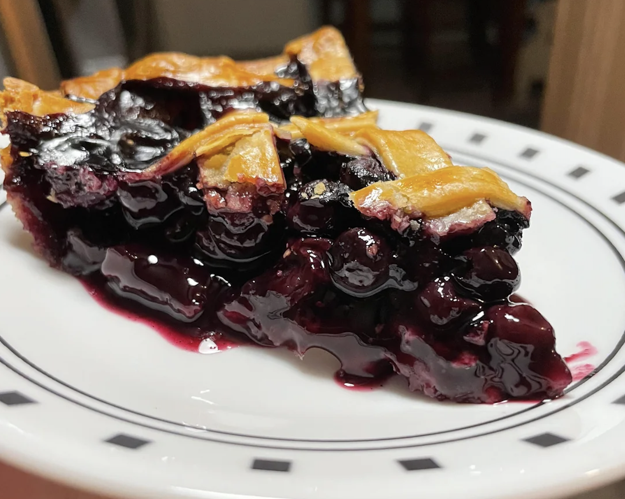 A close-up of a slice of blueberry pie with a golden lattice crust on a white plate with a black pattern. The filling is glossy and purple, with some juices oozing out.