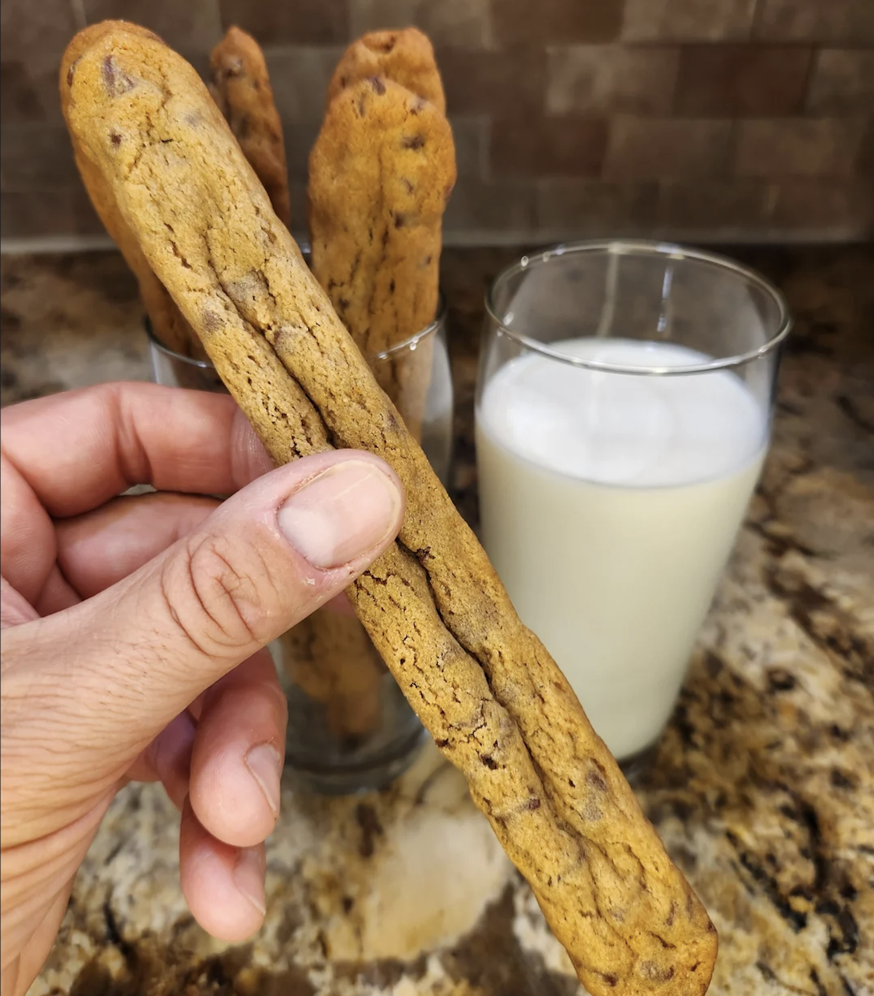 A hand holds a long, thin chocolate chip cookie stick. More cookie sticks are in a glass behind it, next to a glass of milk on a marble countertop.