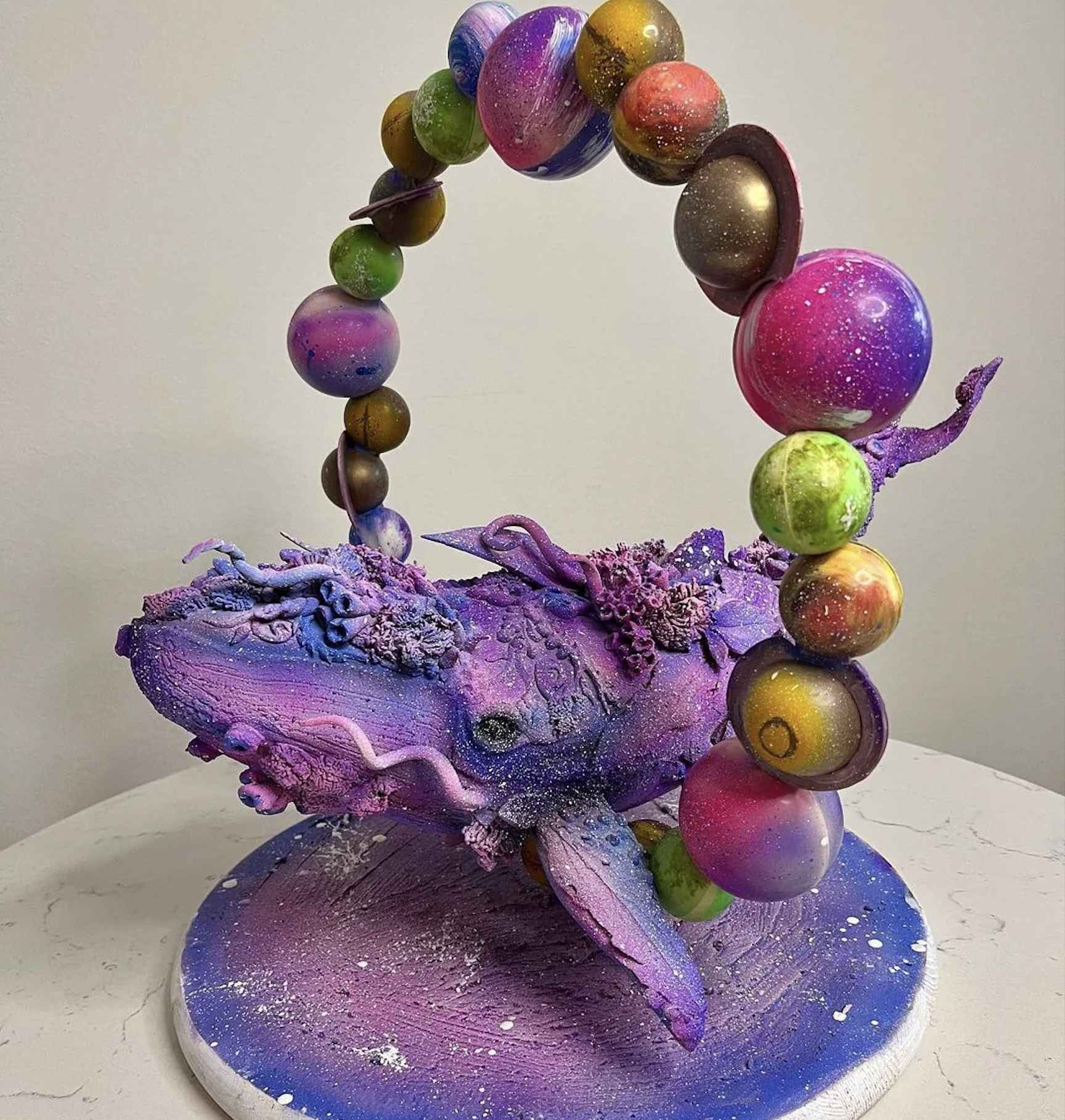 A colorful sculpture of a purple whale with cosmic details. An arch of vibrant, planet-like spheres forms over its back, suggesting a cosmic or fantasy theme. The sculpture sits on a round, starry base.