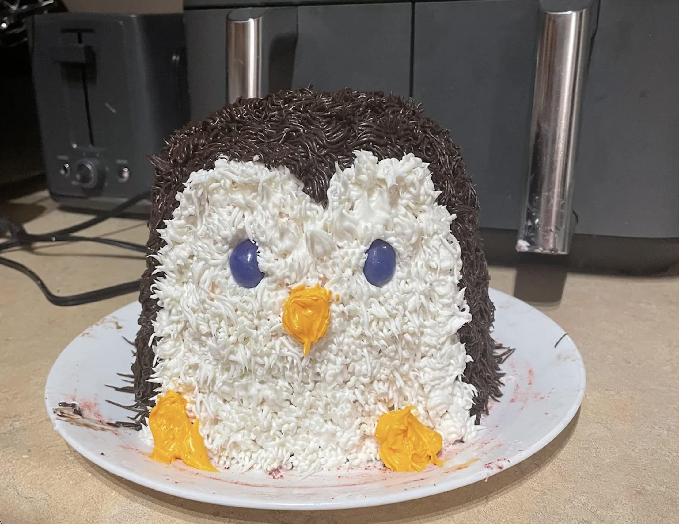 A cake decorated to look like a penguin, with white and dark icing for feathers, blue candy eyes, and orange icing for the beak and feet, sits on a white plate on a kitchen counter.