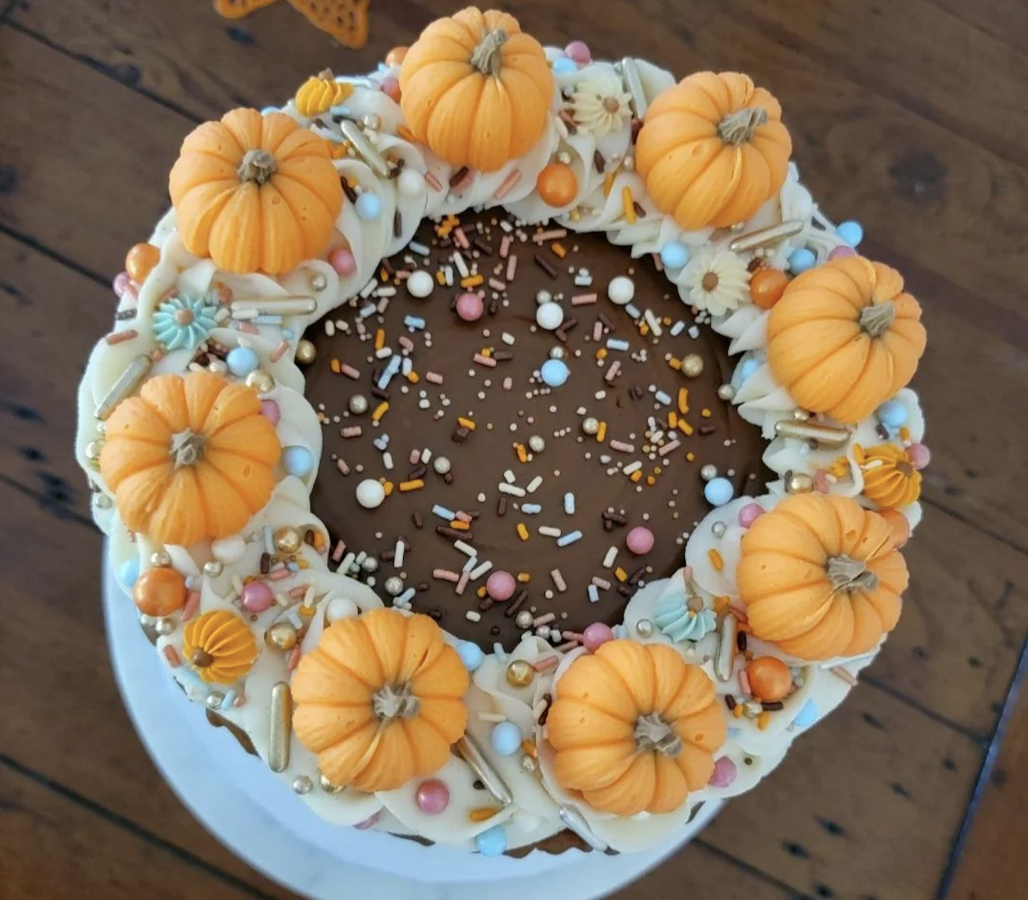 A cake decorated with small orange pumpkin-shaped toppers, colorful sprinkles, and swirled frosting sits on a wooden table.