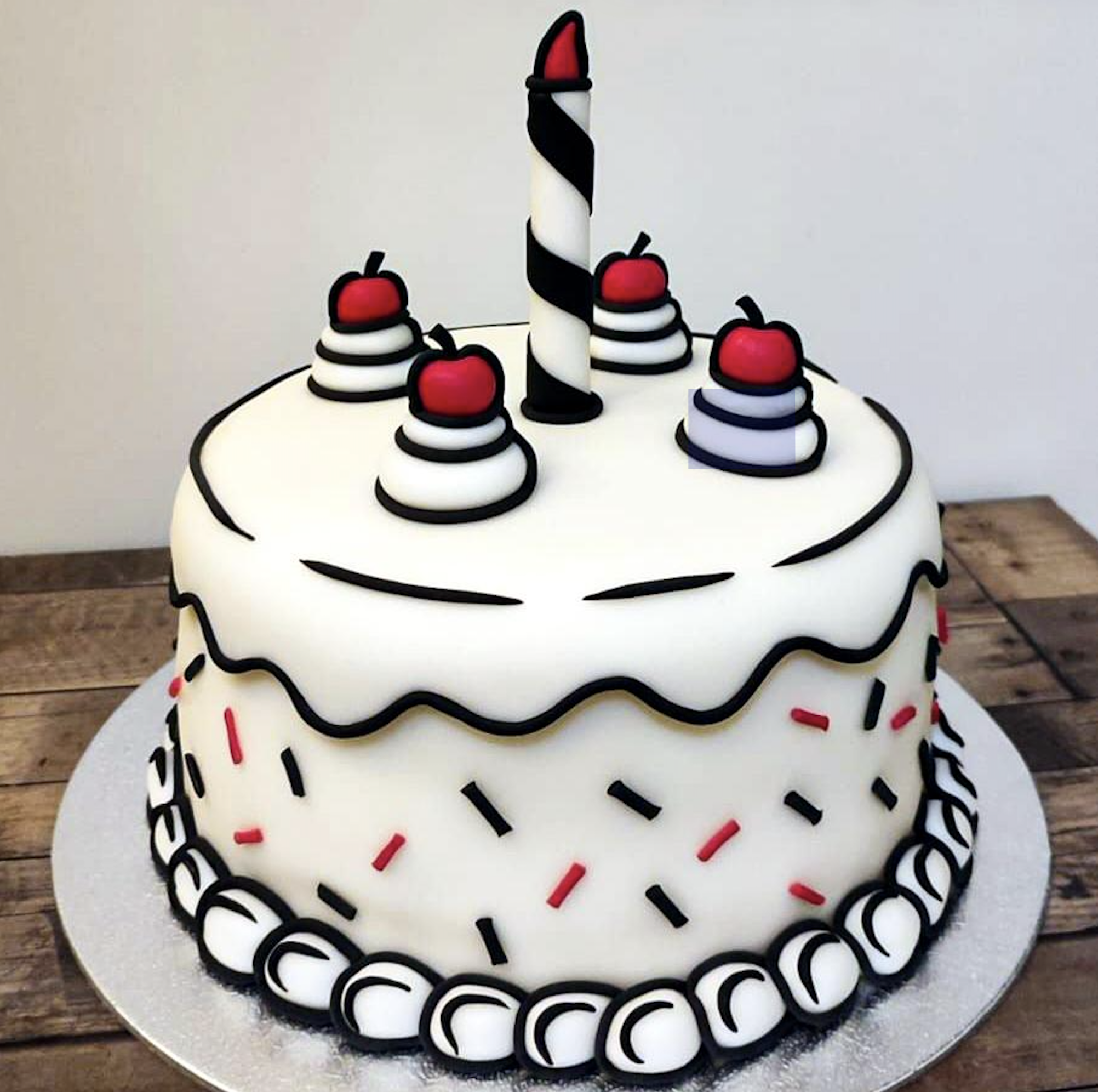 A round, cartoon-style cake with white icing, red and black sprinkles, four cherries on cream swirls, and a single tall candle, sitting on a wooden surface.
