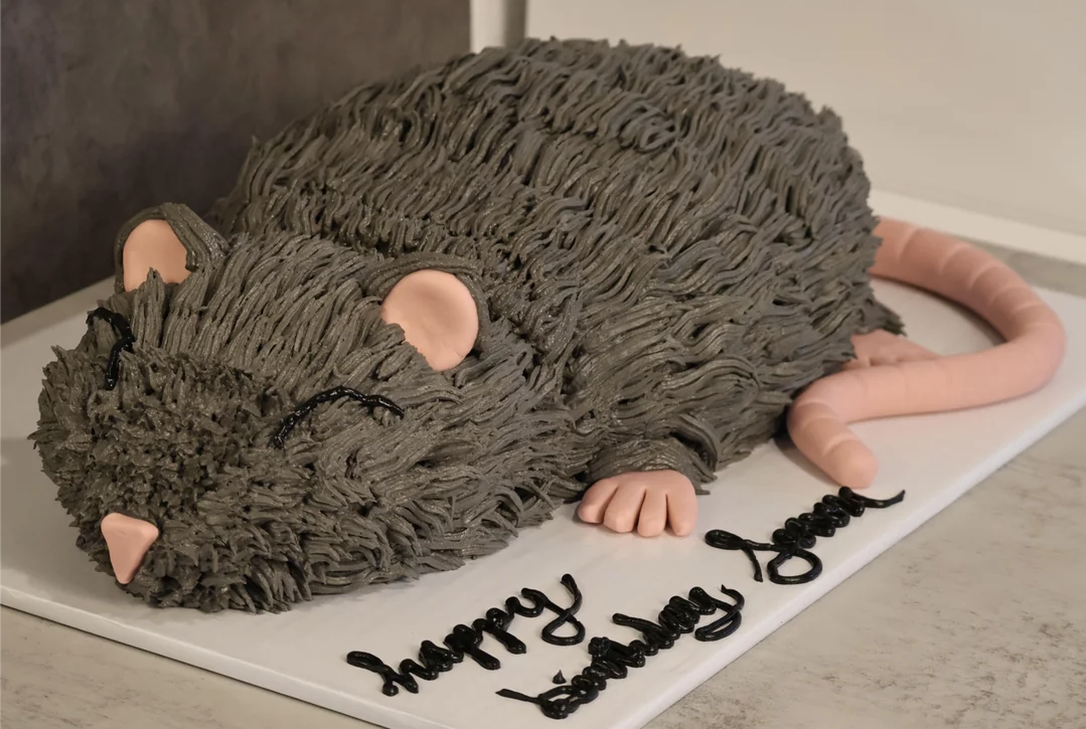 A birthday cake shaped like a gray rat, with pink ears, paws, and tail, resting on a white board that says “Happy Birthday.”