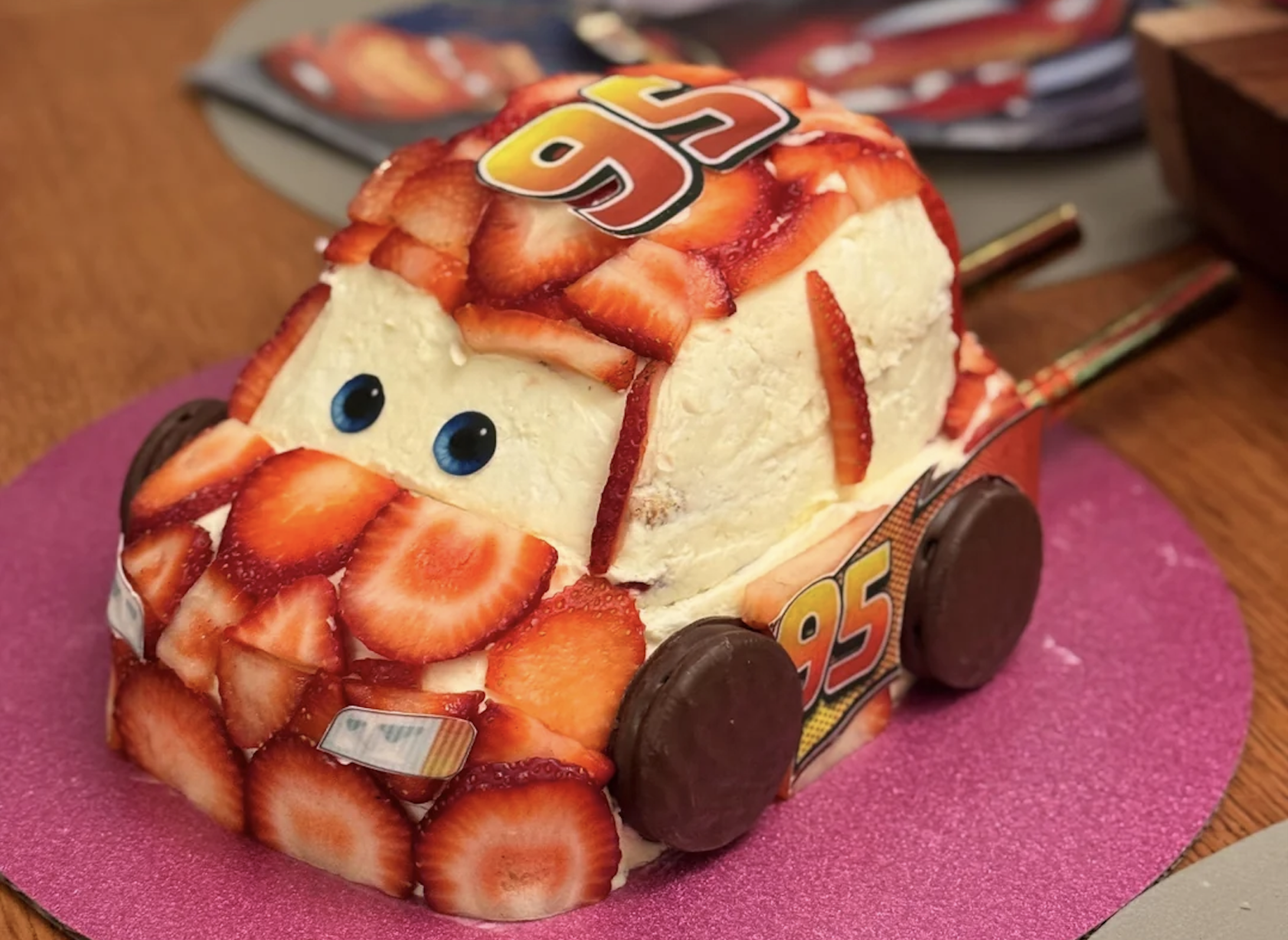 A car-shaped cake decorated with sliced strawberries, chocolate wheels, and the number “95” on top, resembling a character from the movie Cars, sits on a pink mat.
