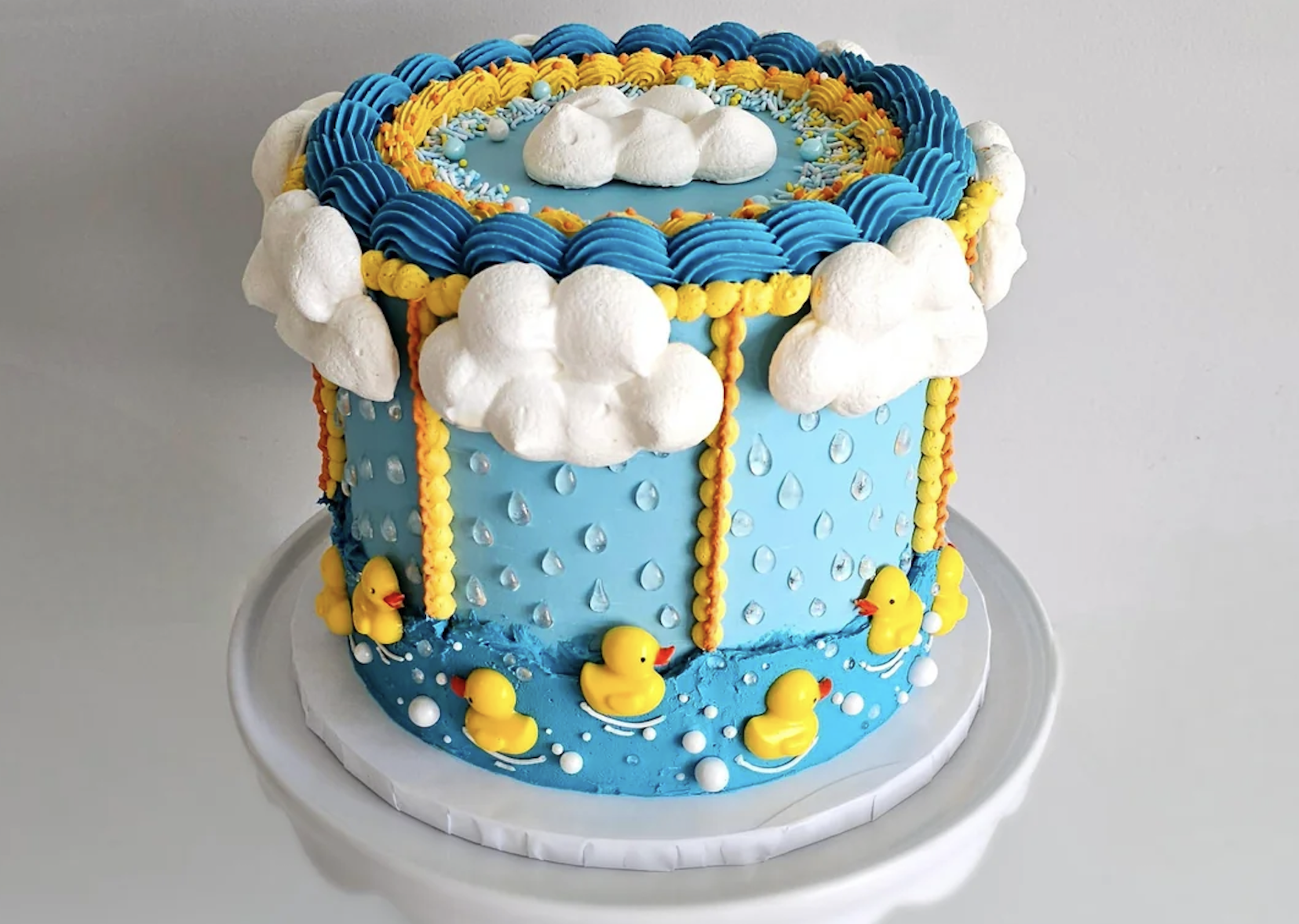A blue cake decorated with white clouds, yellow rain, raindrop details, and small rubber duck figures around the base, resembling a rainy day scene.