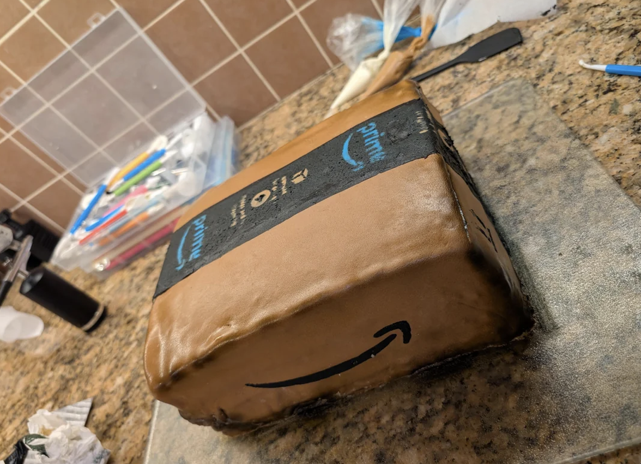 A rectangular cake decorated to look like an Amazon Prime package sits on a granite countertop, with baking tools and supplies in the background.