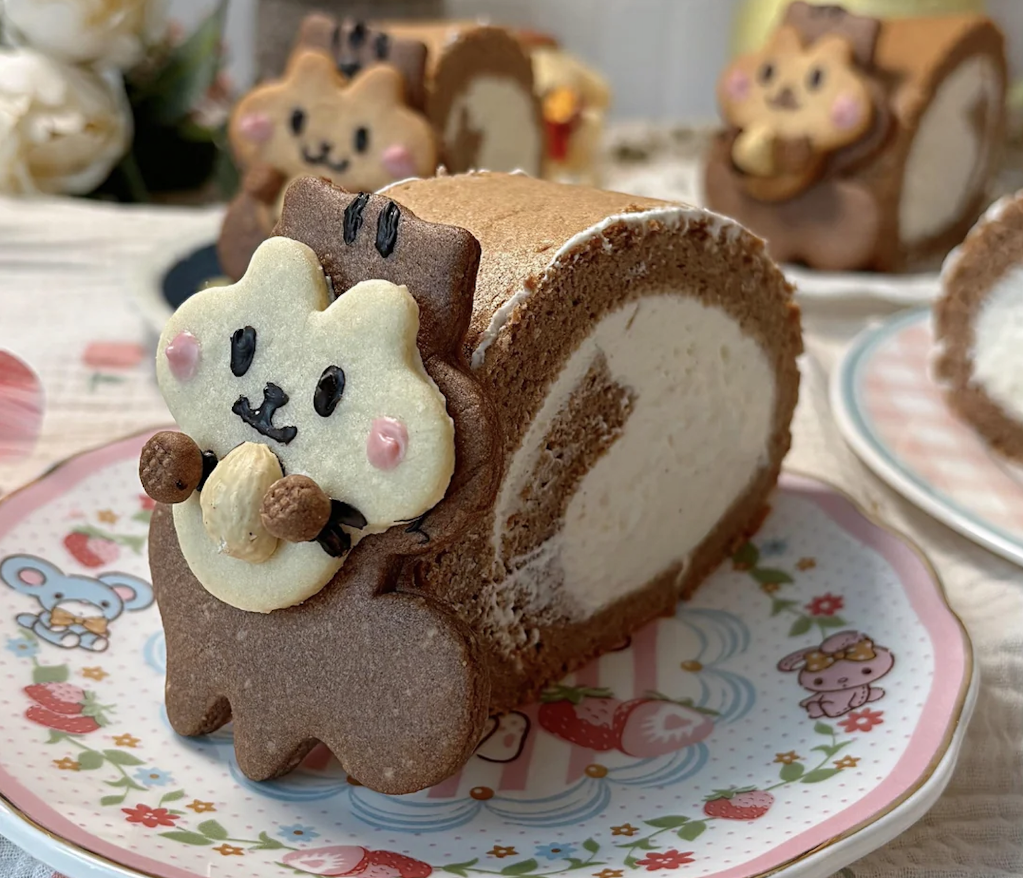 A chocolate and cream Swiss roll is decorated with a cute animal cookie, resembling a squirrel holding a nut, on a pastel plate with cartoon designs. Other similar cakes are in the background.