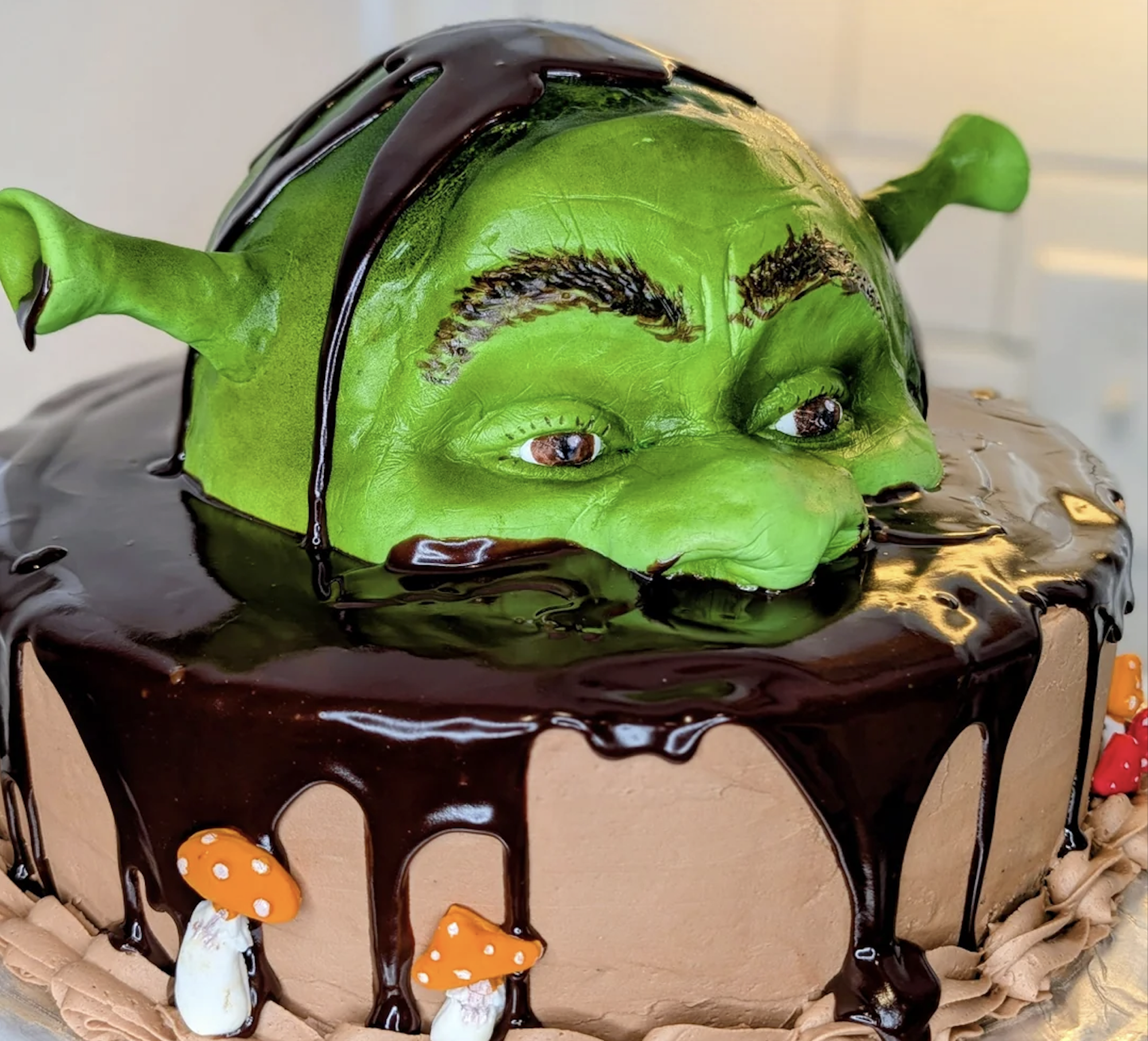 A cake shaped like a green ogre’s head with chocolate icing, resembling Shrek, emerging from a round cake decorated with frosting and small mushroom figurines.