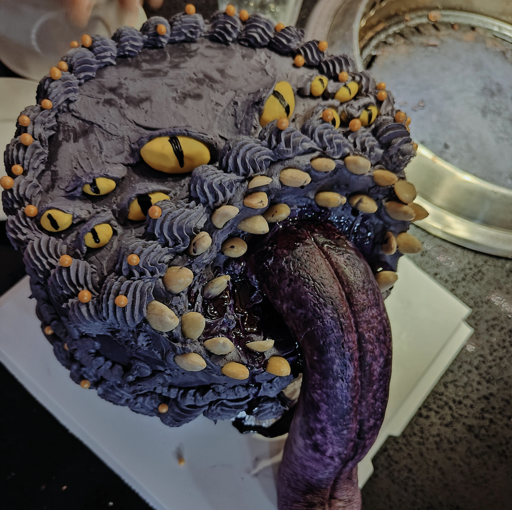 A cake decorated to resemble a monster’s face with yellow eyes, textured purple frosting, and a large, realistic purple tongue extending from its mouth.