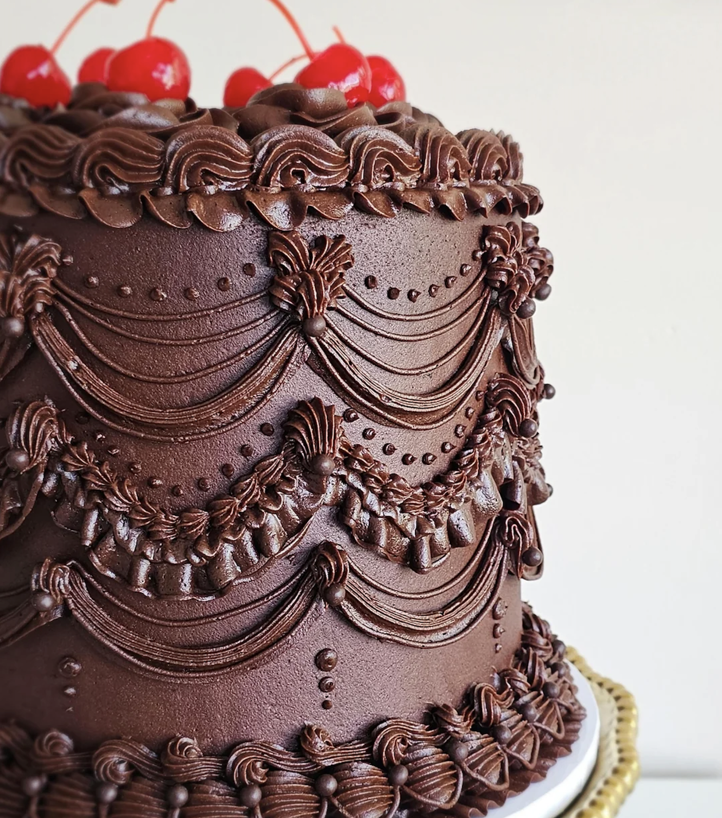 A close-up of a tall, ornate chocolate cake decorated with intricate chocolate icing swags and topped with red cherries, displayed on a gold-rimmed cake stand.