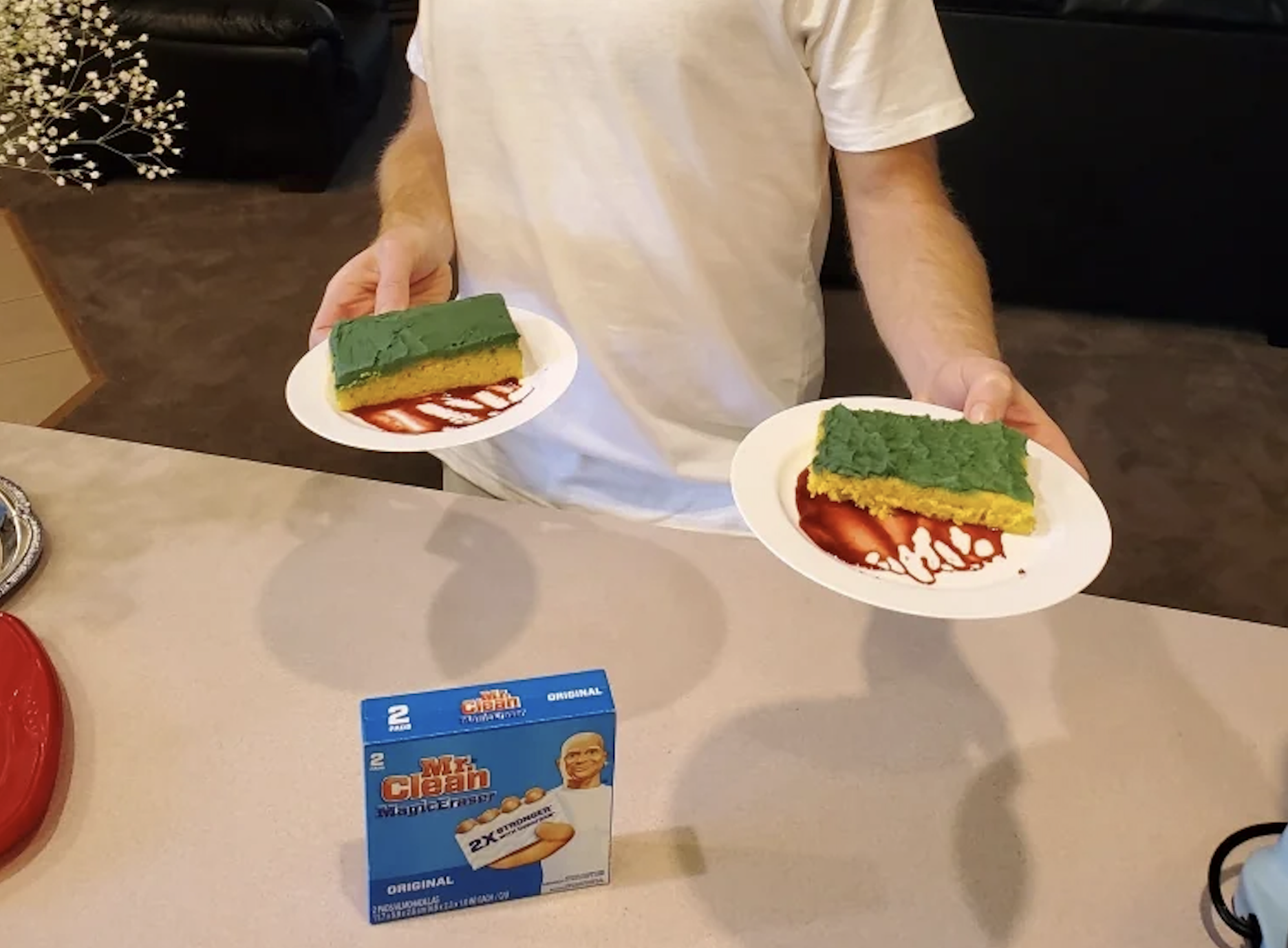 A person holds two plates with yellow-green spongy rectangles on ketchup, resembling dish sponges, with a box of Mr. Clean sponges on the counter.