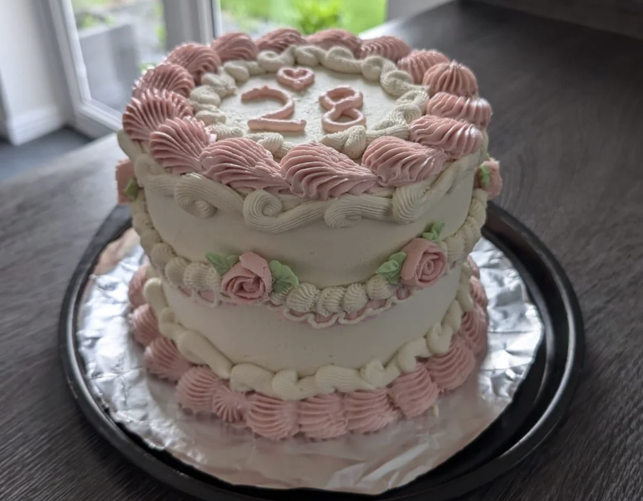 A two-tier cake decorated with pink and white icing, piped swirls, and small roses. The top tier displays the number “28” with a heart. The cake sits on foil and a black tray near a window.