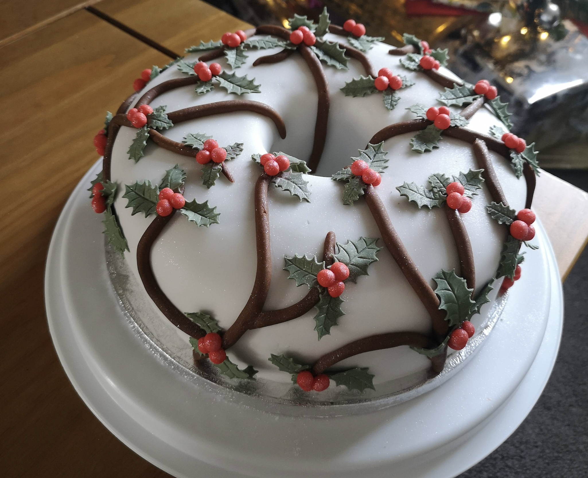 A round cake with white icing, decorated with brown icing vines, green holly leaves, and red berries, sits on a white plate with a Christmas tree blurred in the background.