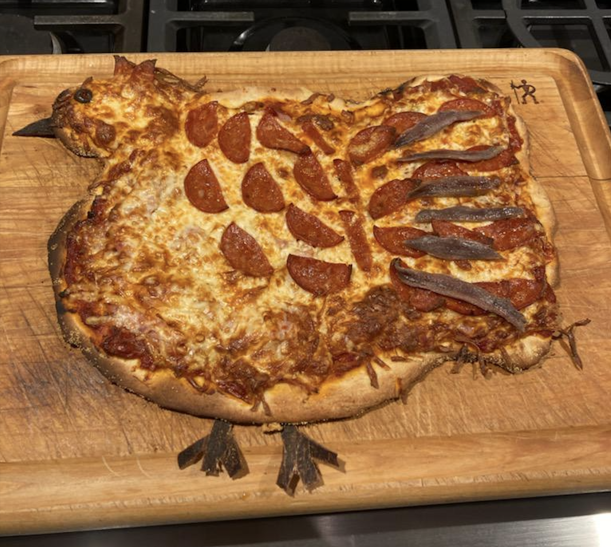 Chicken-shaped pizza with pepperoni and anchovies on a wooden board. Pizza has cheese, pepperoni for feathers, anchovies for wing, and crust shaped to look like a chicken with pepper legs and beak.