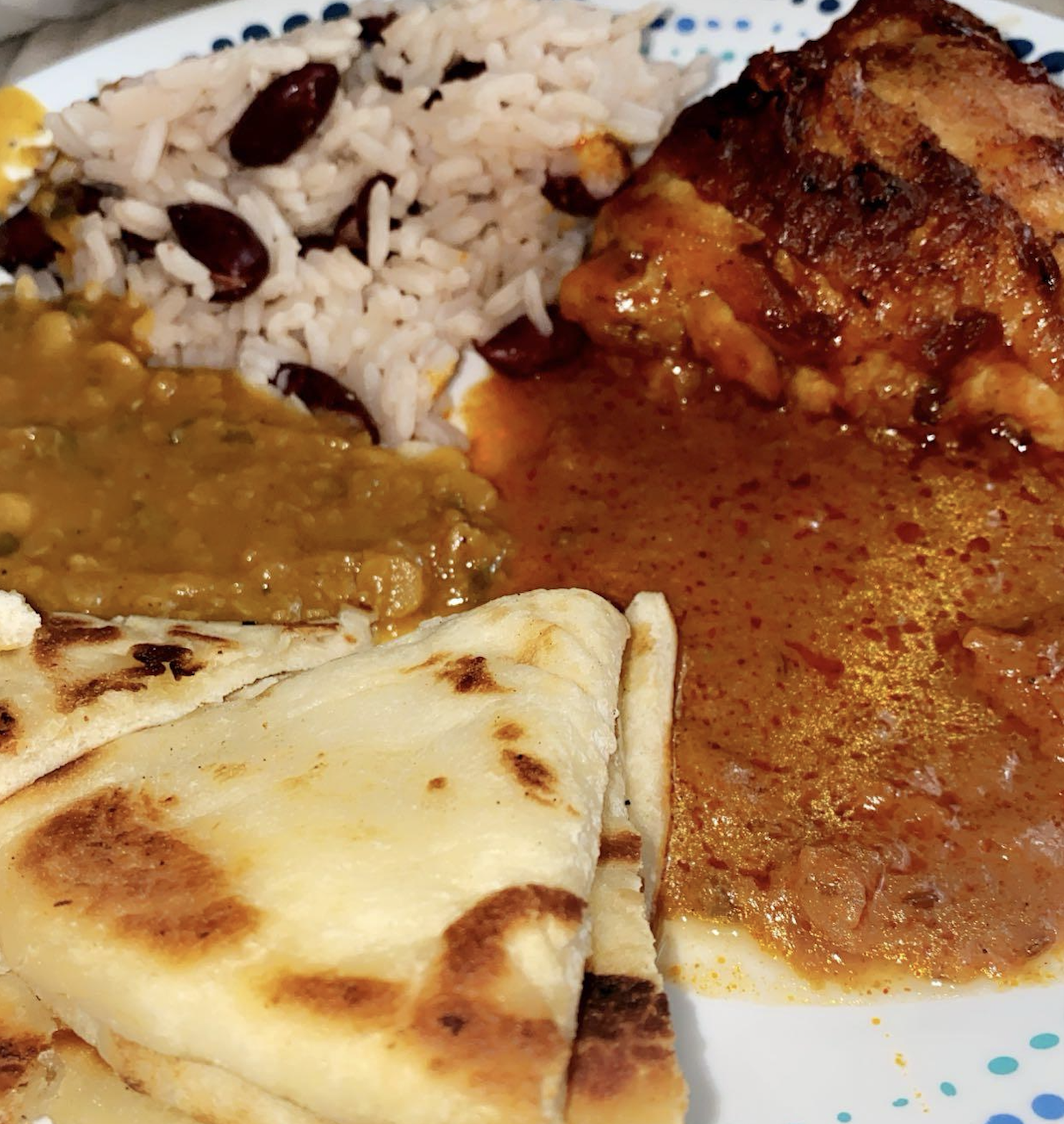 A plate with rice and beans, a piece of stewed chicken in sauce, creamy lentils, and folded pieces of naan bread.