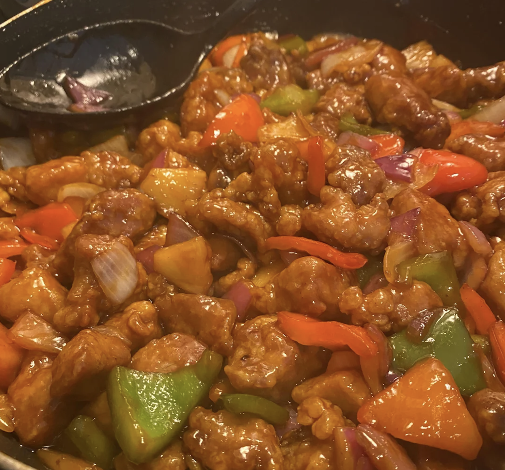 A close-up of sweet and sour pork with chunks of crispy meat, red and green bell peppers, pineapple, and red onions coated in a glossy sauce. A black serving spoon rests on the edge of the dish.