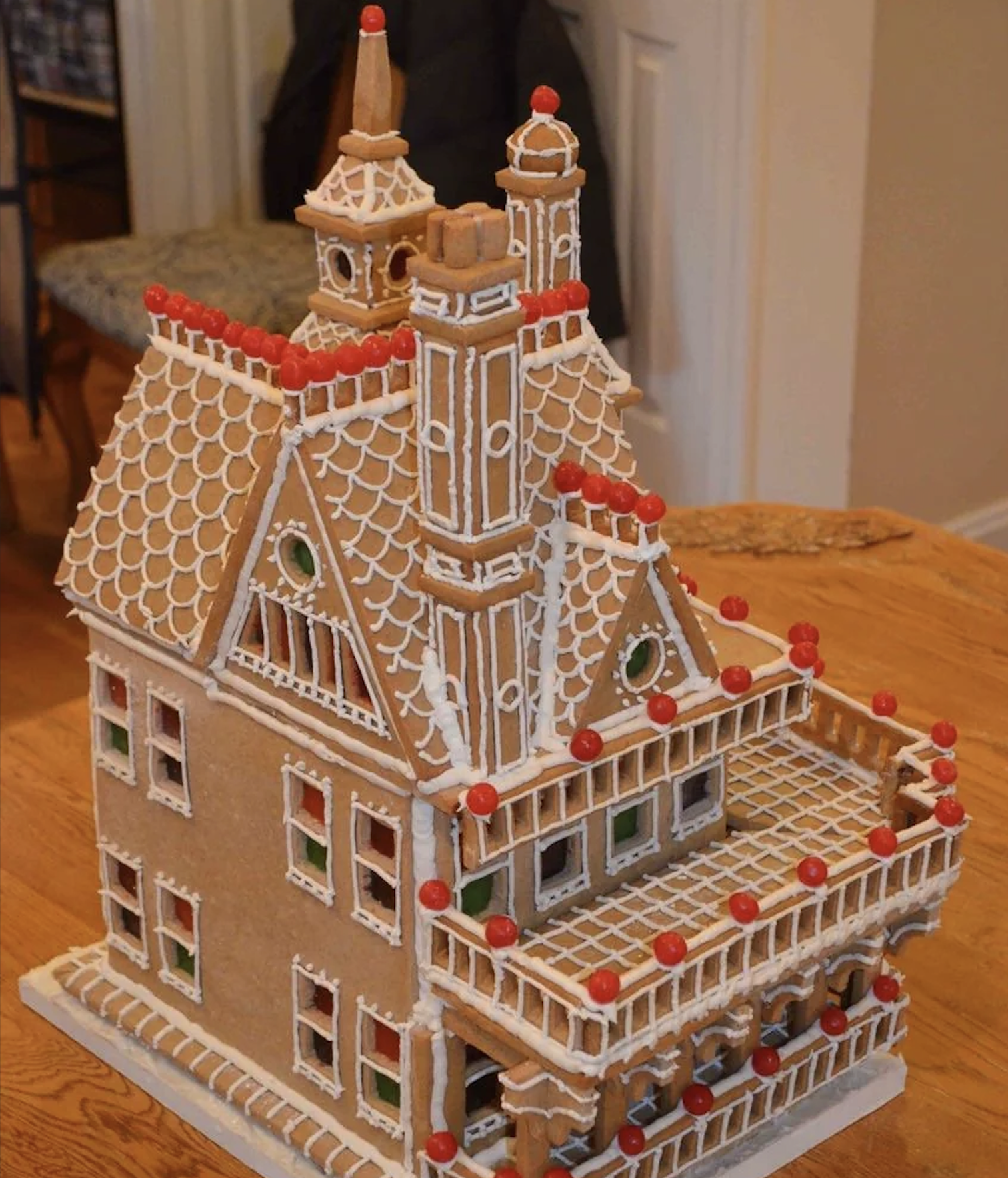 A detailed gingerbread house resembling a Victorian mansion, decorated with white icing and red candy accents, sits on a wooden table indoors.