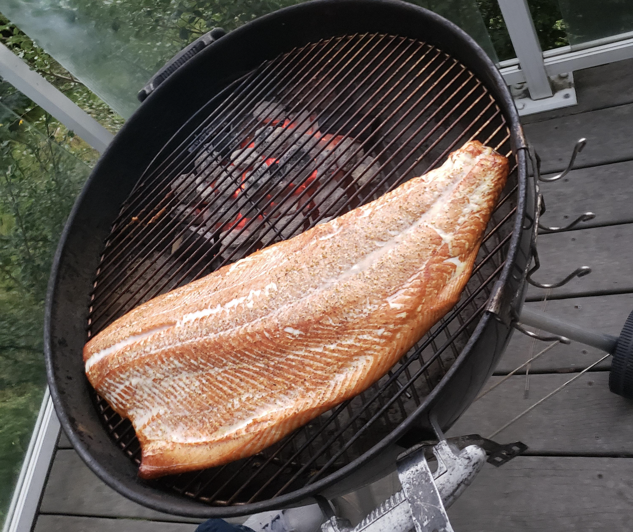 A large, seasoned fillet of fish is grilling over hot coals on a round outdoor charcoal grill, placed on a wooden deck with some greenery visible in the background.